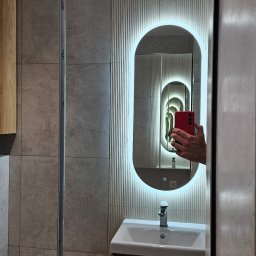 Daniel Poczobut DP Projekt Detal - Nowoczesna łazienka z podświetlanym lustrem owalnym, umywalką na szafce z drewnianym frontem i szarą płytką na ścianie. W lustrze odbija się telefon.