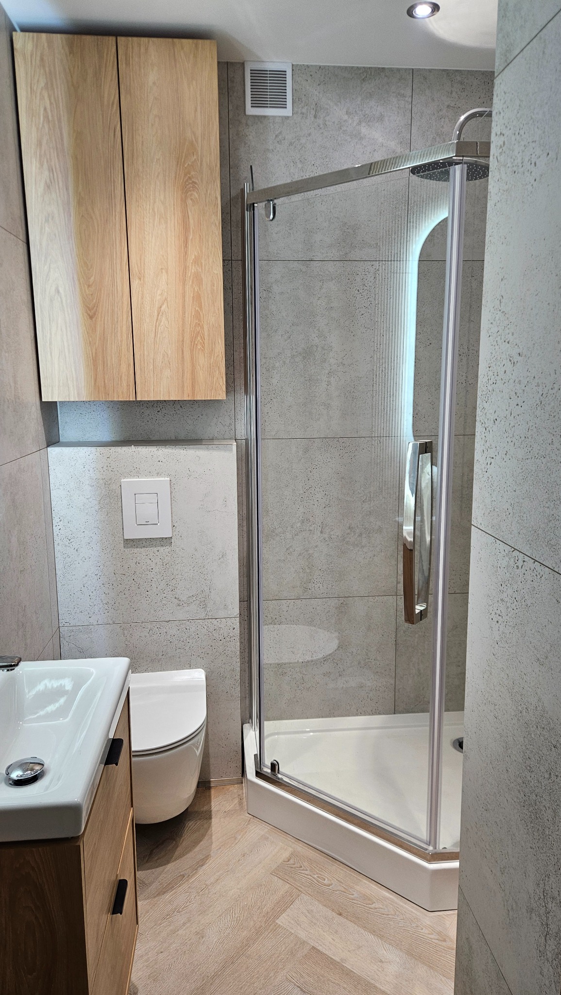 Nowoczesna łazienka z kabiną prysznicową, umywalką z szafką z drewna i toaletą. Szare płytki na ścianach i podłodze imitują naturalny kamień. Minimalistyczny design.