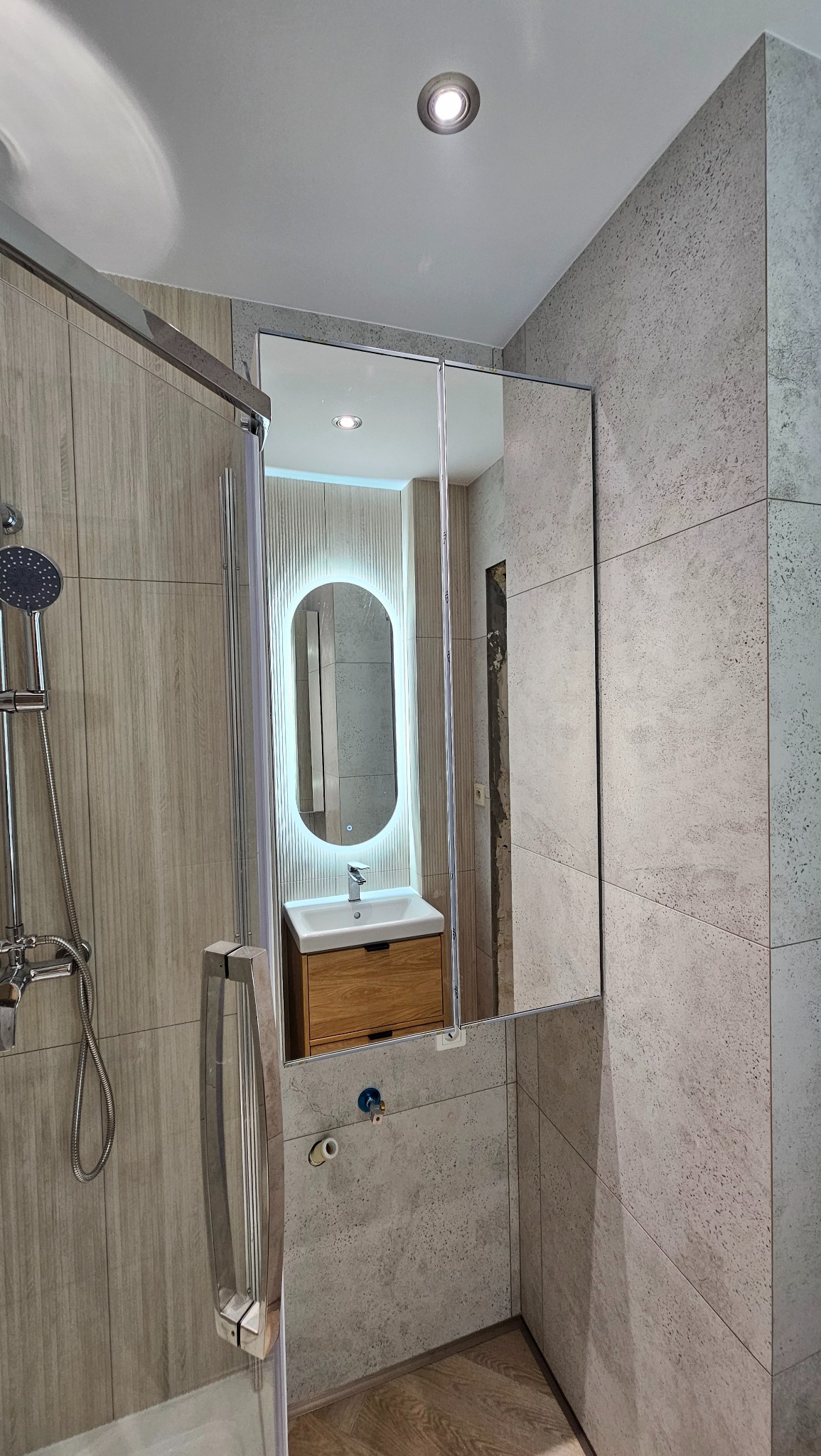 Nowoczesna łazienka z kabiną prysznicową, lustrem z podświetleniem LED i drewnianą szafką pod umywalką. Szare płytki imitujące kamień na ścianach i podłodze.