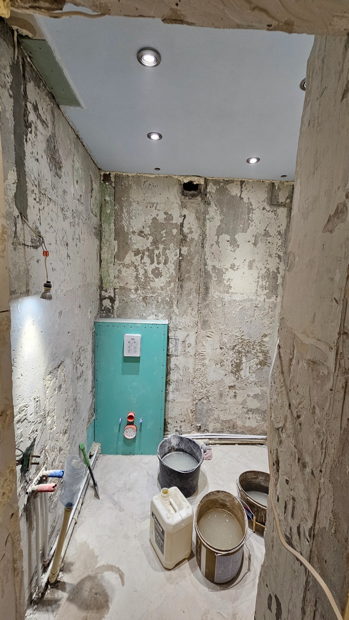 Łazienka w trakcie remontu: surowe ściany z widocznymi warstwami, instalacja podtynkowa, wiadra z zaprawą i narzędzia. Oświetlenie sufitowe punktowe.