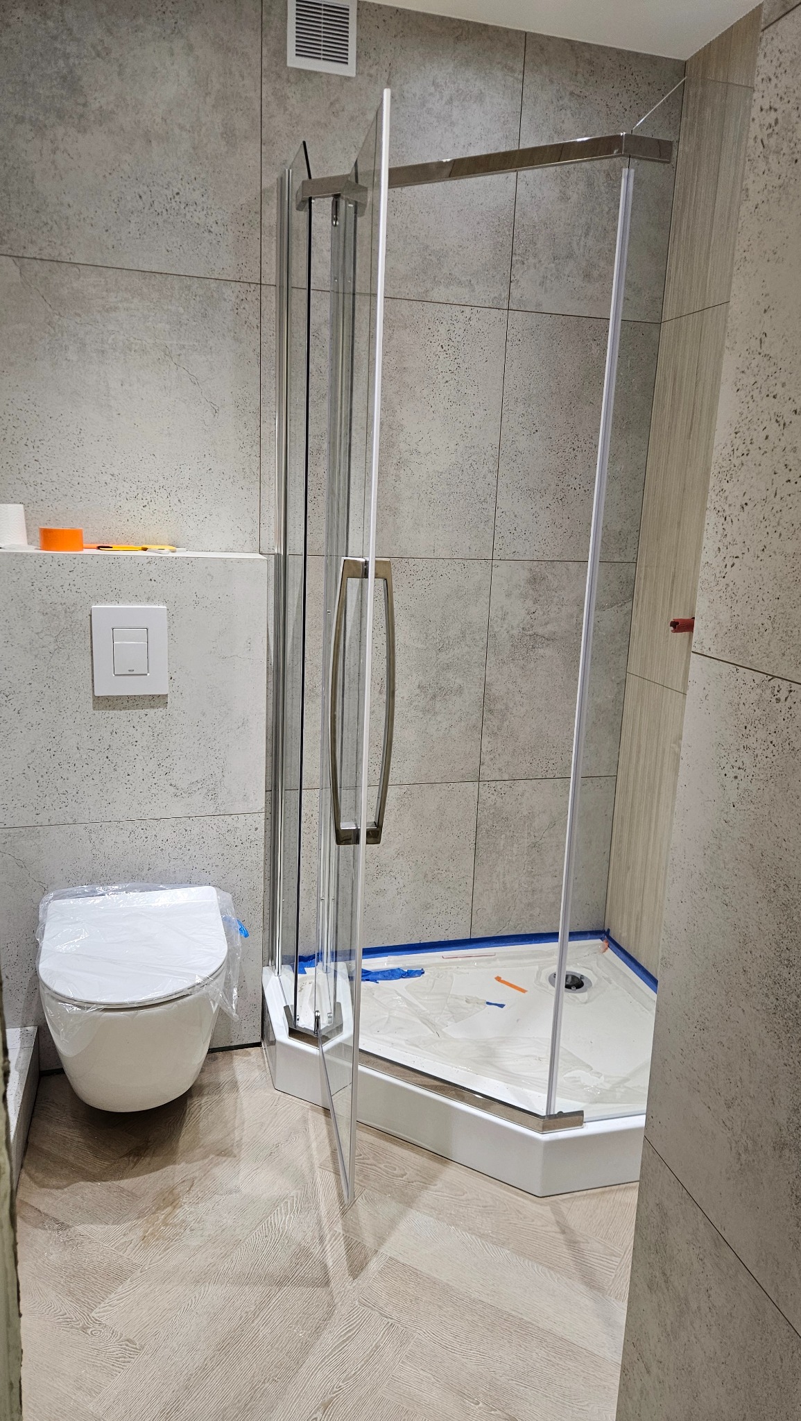 Nowoczesna łazienka w trakcie wykańczania: kabina prysznicowa z otwartymi drzwiami, toaleta z folią ochronną i szare płytki imitujące beton na ścianach.