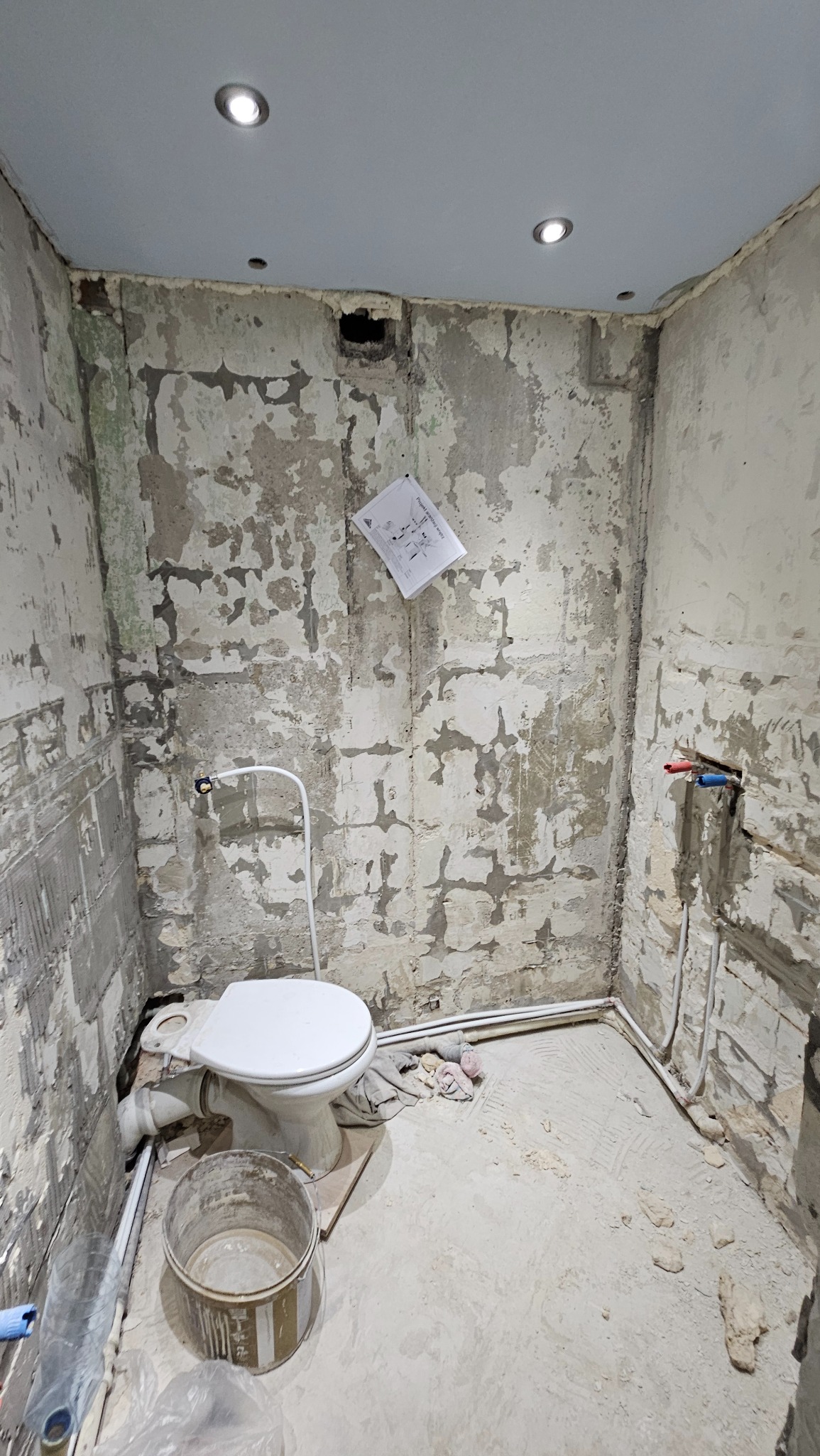 Łazienka w trakcie remontu: ściany ze śladami starej glazury, wiszący schemat instalacji, częściowo zamontowana toaleta i wiadro. Widoczne instalacje wodne.