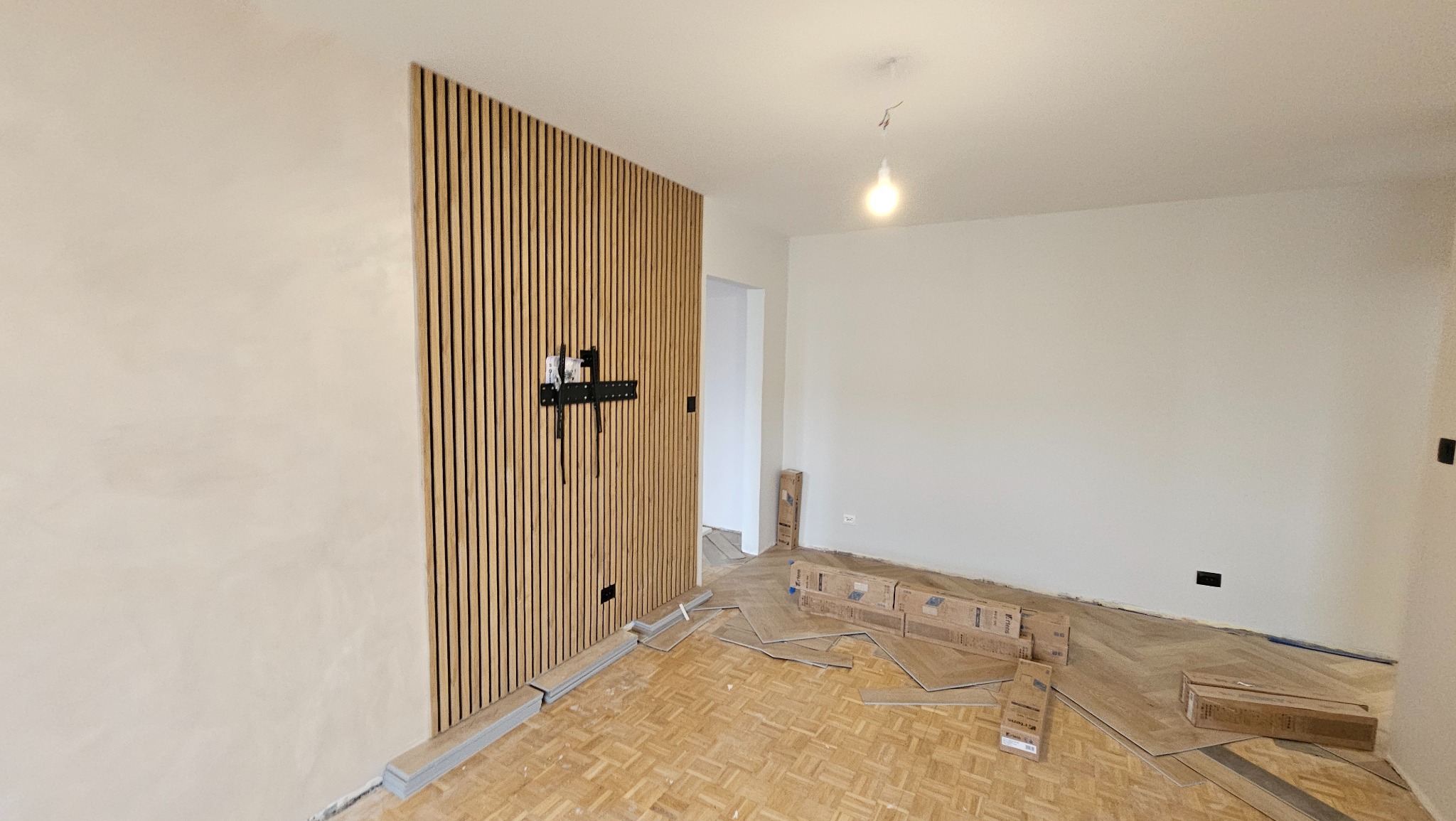 Pokój w trakcie remontu: drewniana lamelowa ściana z uchwytem TV, parkiet w jodełkę, kartony z panelami podłogowymi. Widoczne gniazdka elektryczne.