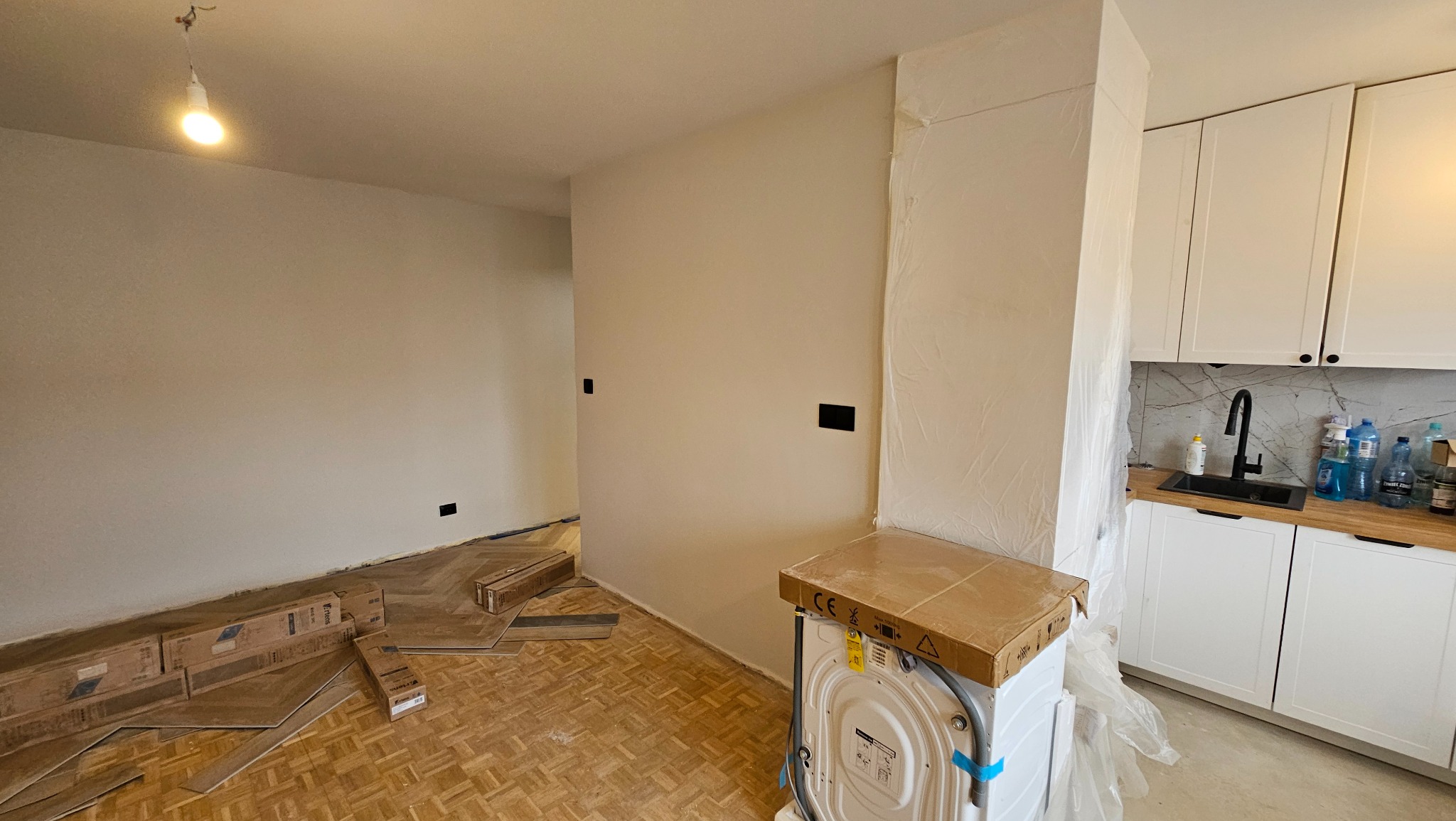 Wnętrze w trakcie remontu: ułożony parkiet, ściany przygotowane do malowania, zabudowa kuchenna z białymi szafkami i czarną armaturą. Widoczne materiały budowlane.