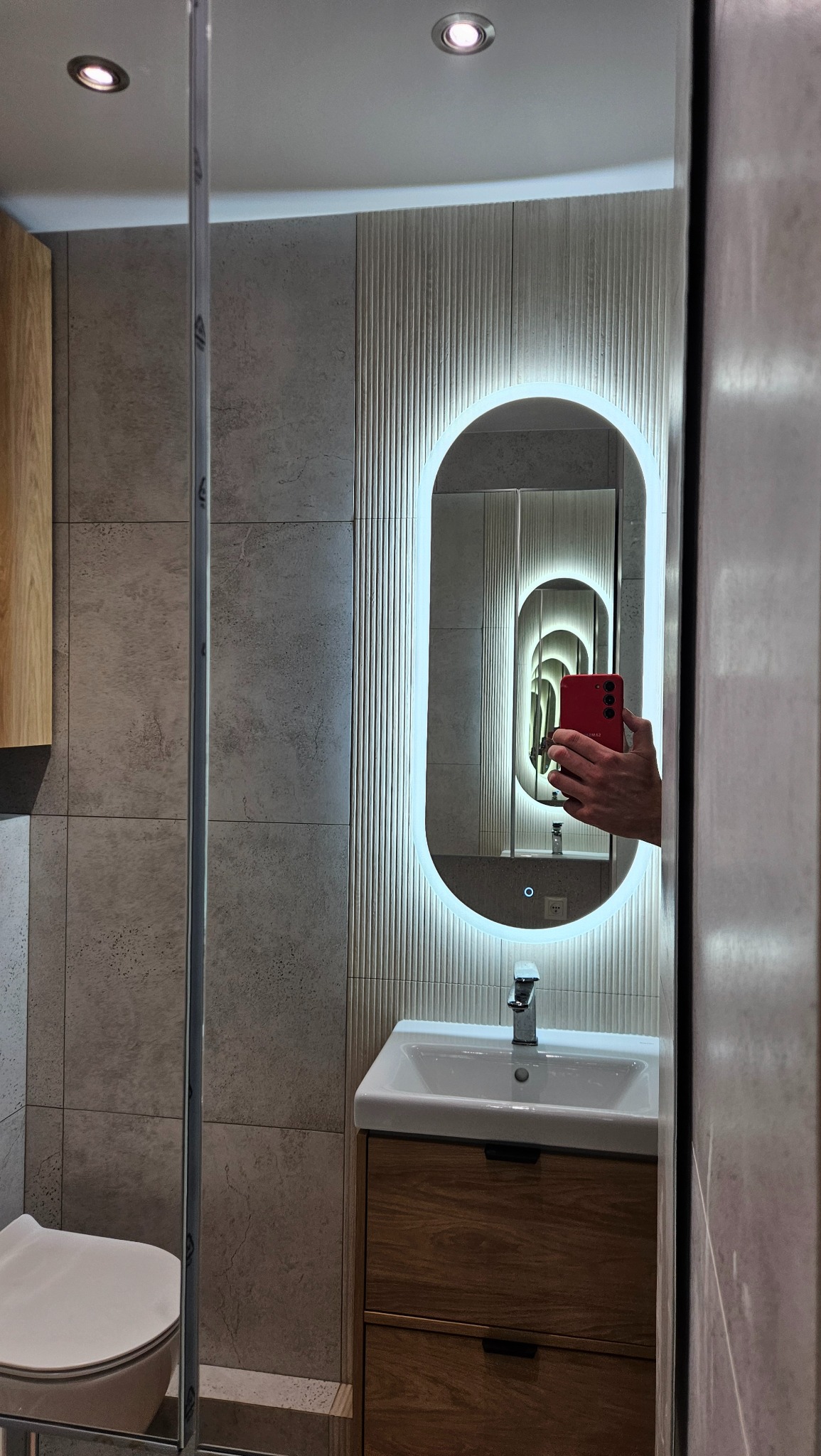 Nowoczesna łazienka z podświetlanym lustrem owalnym, umywalką na szafce z drewnianym frontem i szarą płytką na ścianie. W lustrze odbija się telefon.