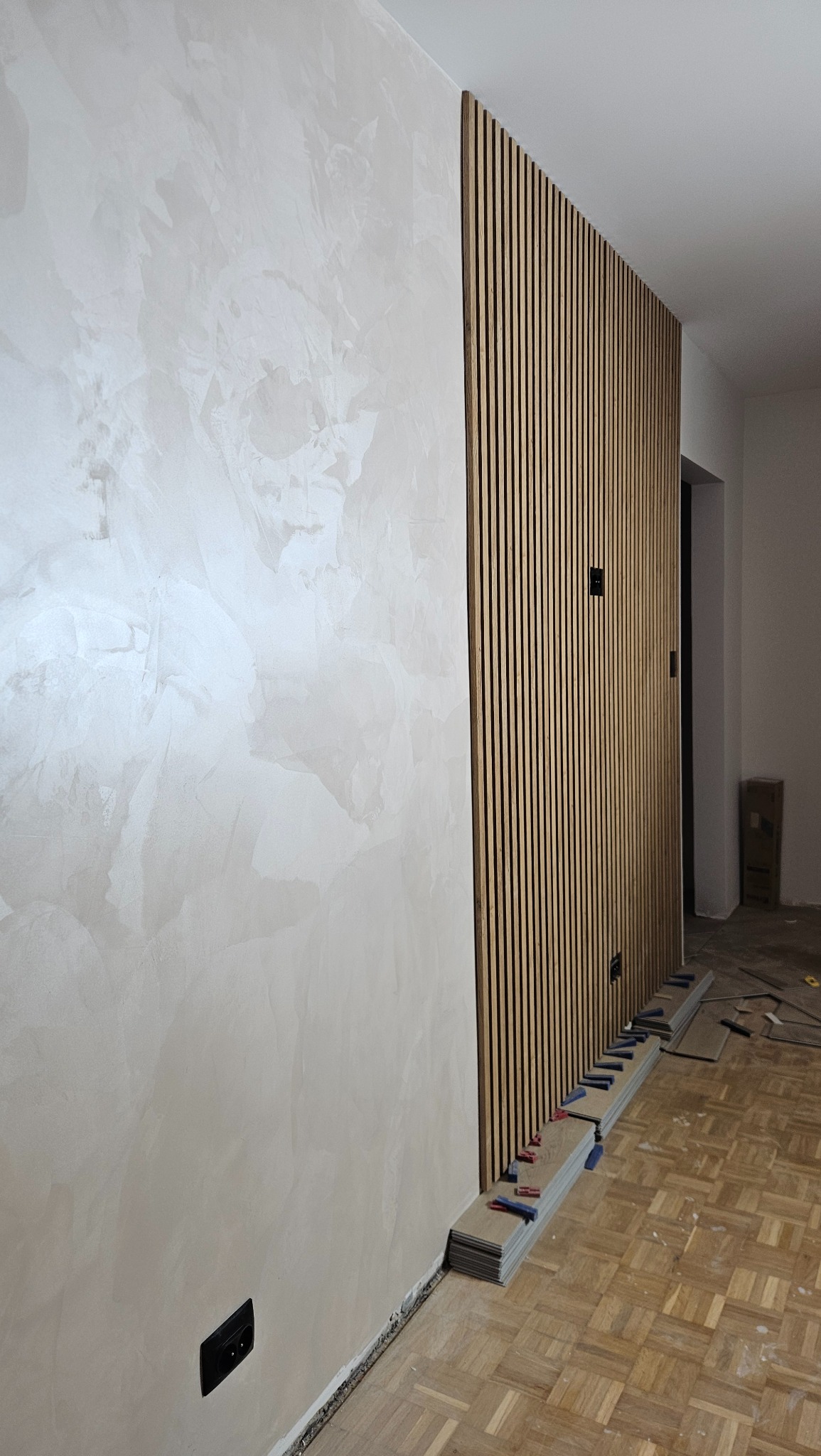 Ściana z dekoracyjną strukturą i drewnianymi lamelami. Widoczne narzędzia i materiały na podłodze. Ujęcie z bliska, prezentujące detale wykończenia wnętrza.