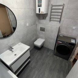 Firma sprzątająca - Nowoczesna łazienka w szarościach: umywalka z szafką, toaleta podwieszana, grzejnik drabinkowy i pralka. Minimalistyczny design, płytki imitujące beton.