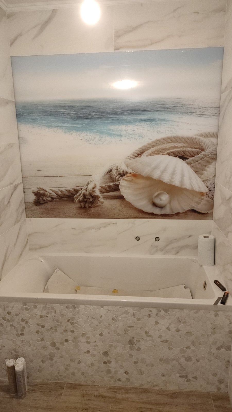 Wykończona łazienka z wanną, mozaiką i dekoracyjnym panelem ściennym z motywem morskim, ukazującym muszlę i linę na tle plaży. Widoczne narzędzia i materiały wykończeniowe.