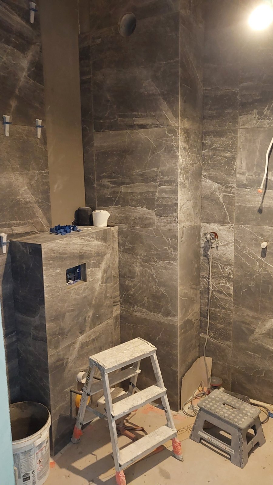 Łazienka w trakcie wykańczania: szare płytki imitujące marmur na ścianach, drabina malarska, widoczne instalacje. Etap prac wykończeniowych.