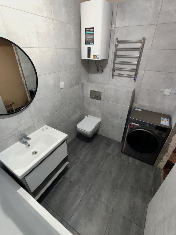 Nowoczesna łazienka w szarościach: umywalka z szafką, toaleta podwieszana, grzejnik drabinkowy i pralka. Minimalistyczny design, płytki imitujące beton.