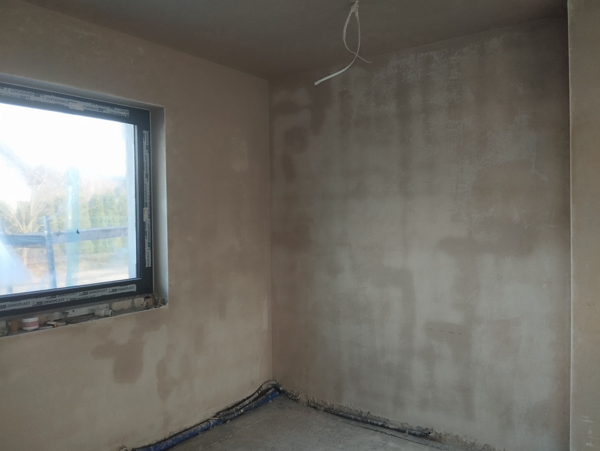 Świeżo nałożone, surowe tynki na ścianach w rogu pokoju z oknem. Widoczne przewody przy podłodze i kabel zwisający z sufitu. Tynk w odcieniach beżu i szarości.