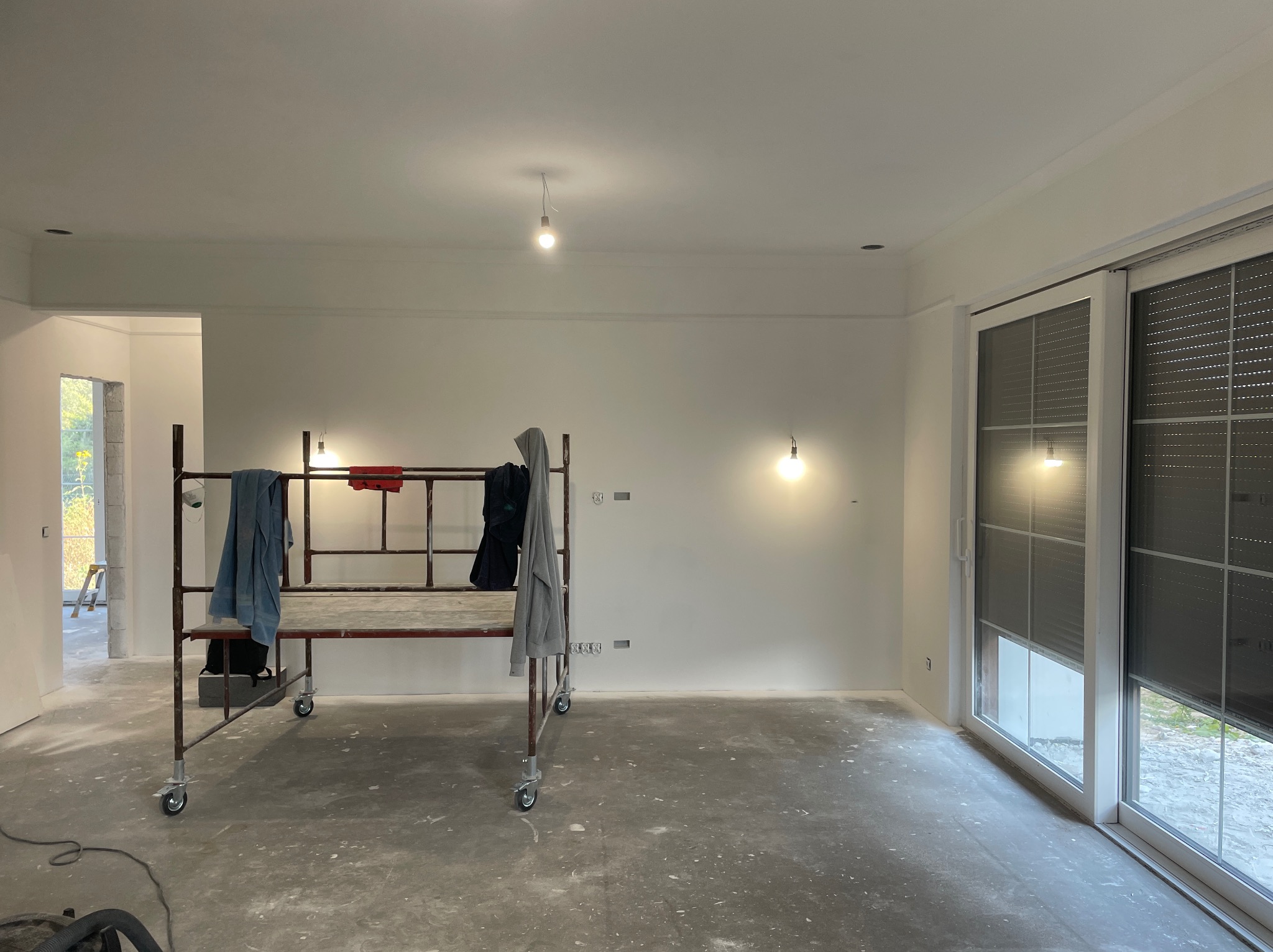 Wnętrze w trakcie prac wykończeniowych: pomalowane ściany, surowa podłoga, rusztowanie z ubraniami, okno przesuwne z roletami. Widoczne punkty oświetleniowe.