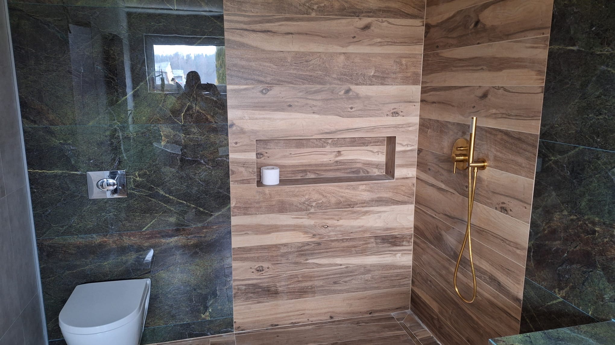 Nowoczesna łazienka z płytkami imitującymi drewno i zielonym marmurem. Złota armatura prysznicowa i wnęka z rolką papieru toaletowego. Widoczna toaleta i odbicie w szybie.