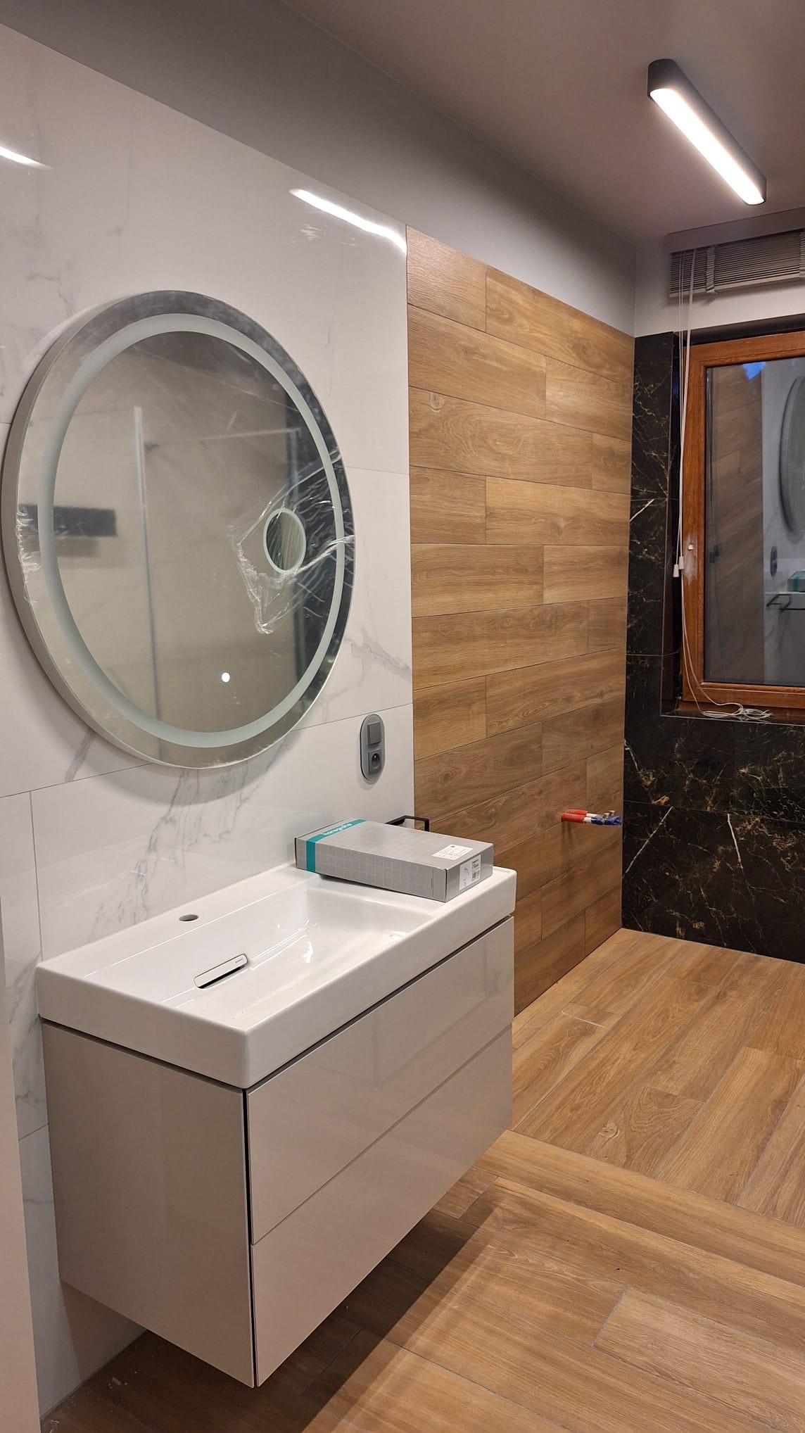 Nowoczesna łazienka po remoncie: biała umywalka z szafką, okrągłe lustro z podświetleniem, płytki imitujące drewno i marmur, widoczne elementy instalacji.