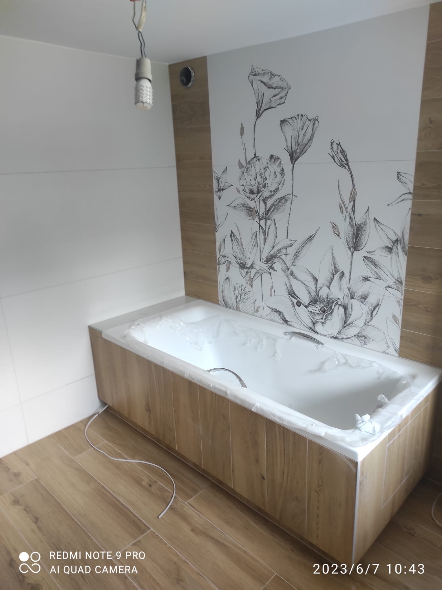 Wykończona łazienka z wanną obudowaną drewnopodobnymi panelami, białe płytki i dekoracyjny motyw kwiatowy na ścianie. Widoczne podłączenie oświetlenia sufitowego.