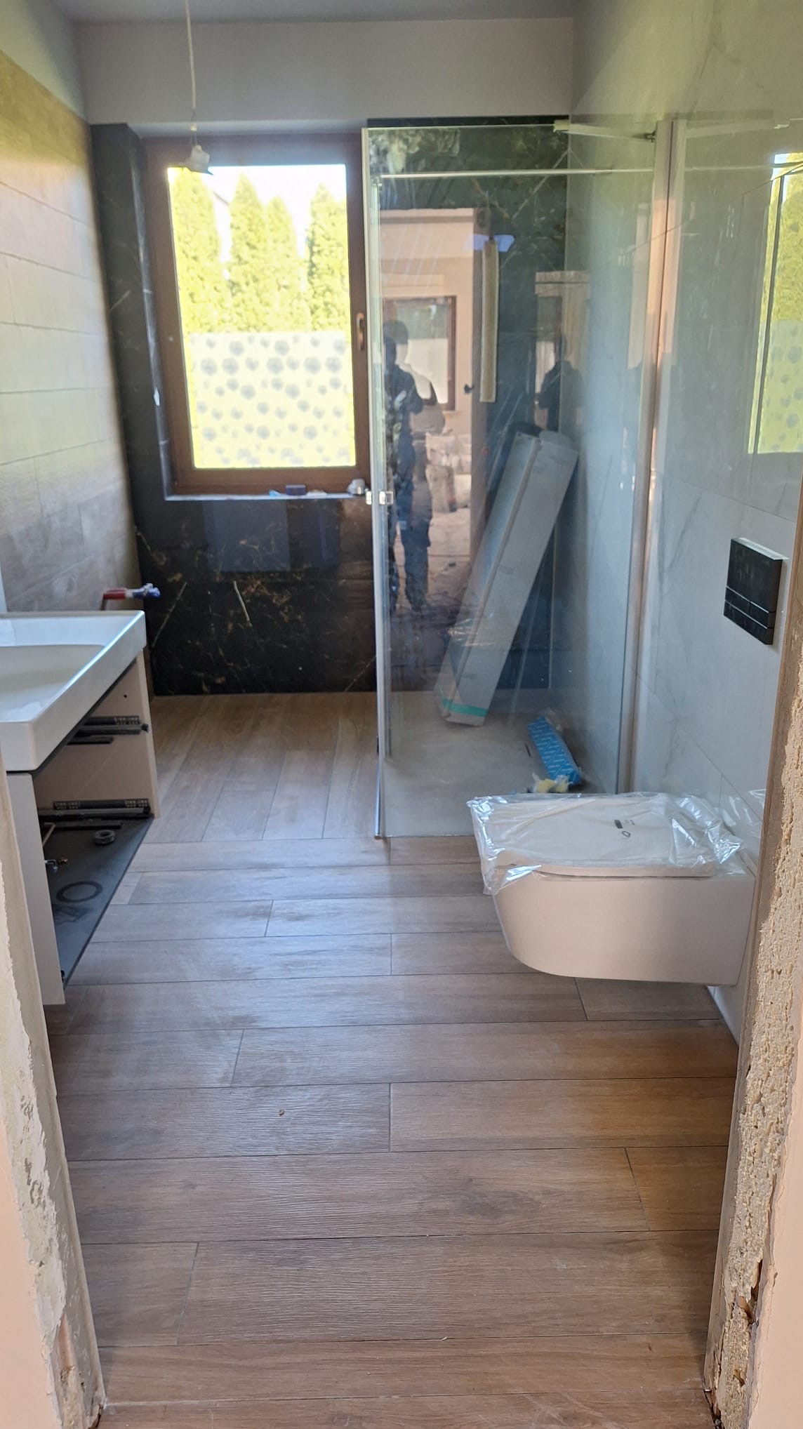 Nowoczesna łazienka w trakcie wykańczania: drewnopodobna podłoga, kabina prysznicowa ze szkła, podwieszana toaleta z folią ochronną, okno z widokiem na zieleń.