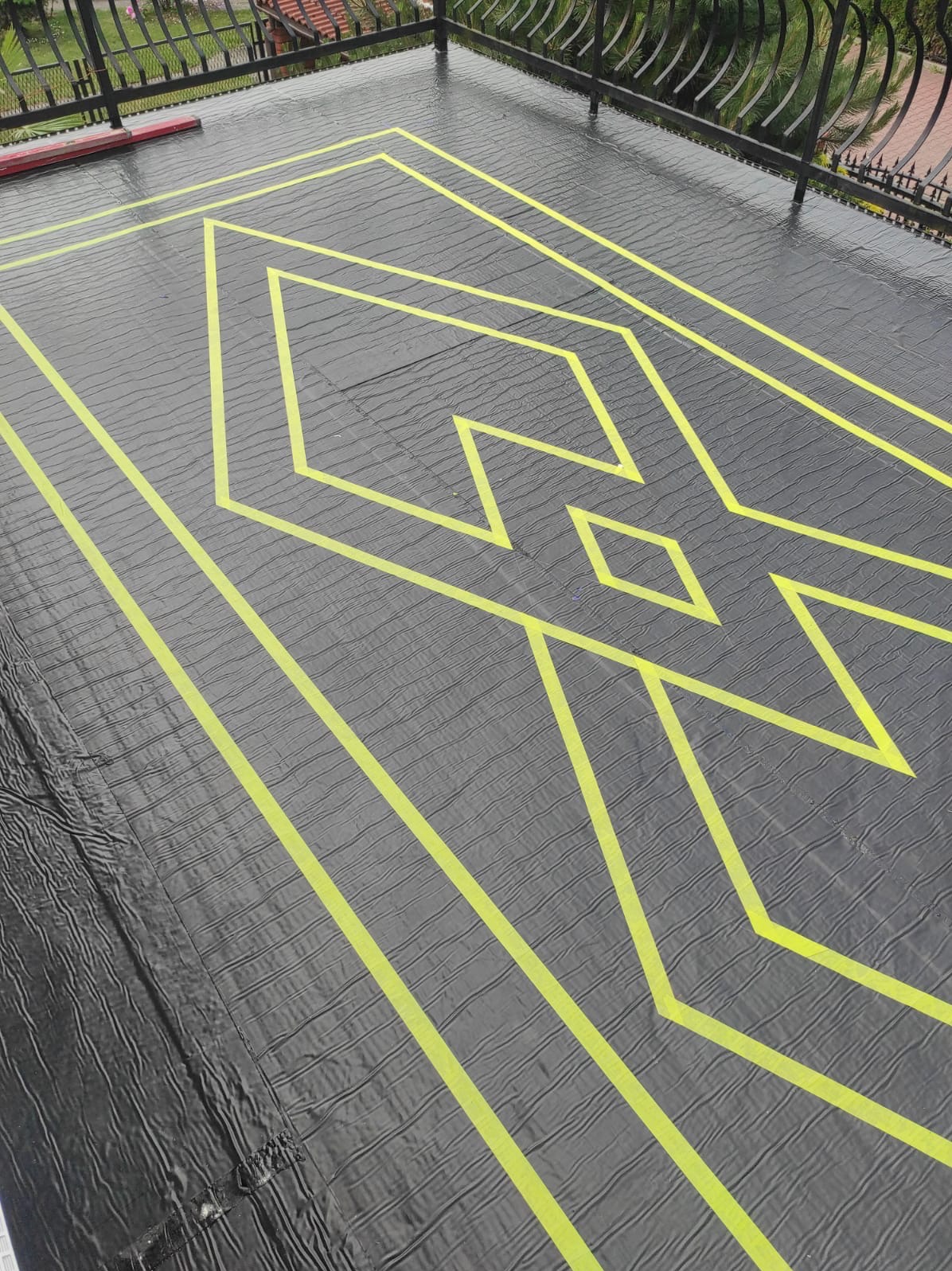 Czarna, błyszcząca powierzchnia balkonu z geometrycznym wzorem wyklejonym żółtą taśmą malarską. Widok z góry, balustrada w tle.