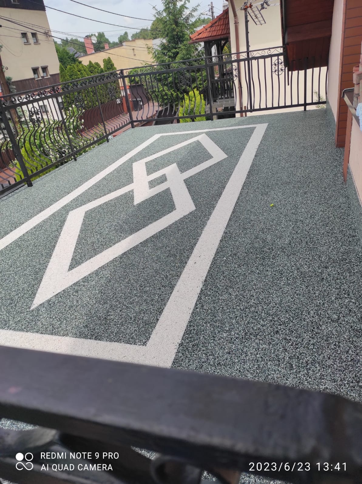 Balkon z szarą, kamyczkową posadzką i białym, geometrycznym wzorem. Metalowa balustrada. Widok na budynki i zieleń w tle.