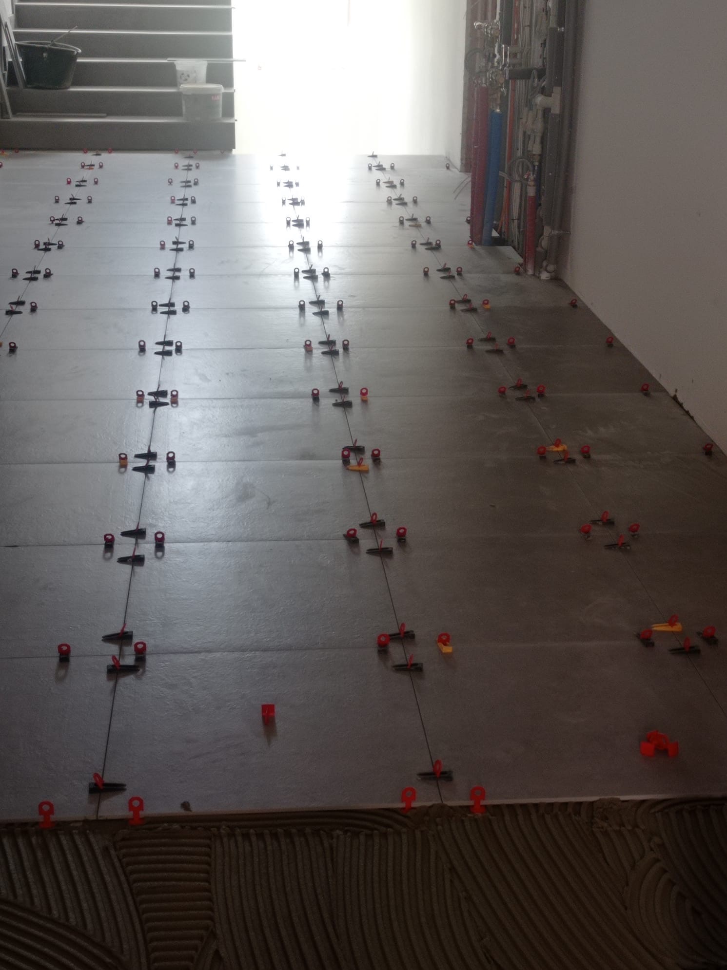 Szare płytki podłogowe w trakcie układania, z systemem poziomowania (czerwone klipsy i kliny). Widoczna warstwa kleju i fragment schodów w tle. Jasne światło wpadające z góry.