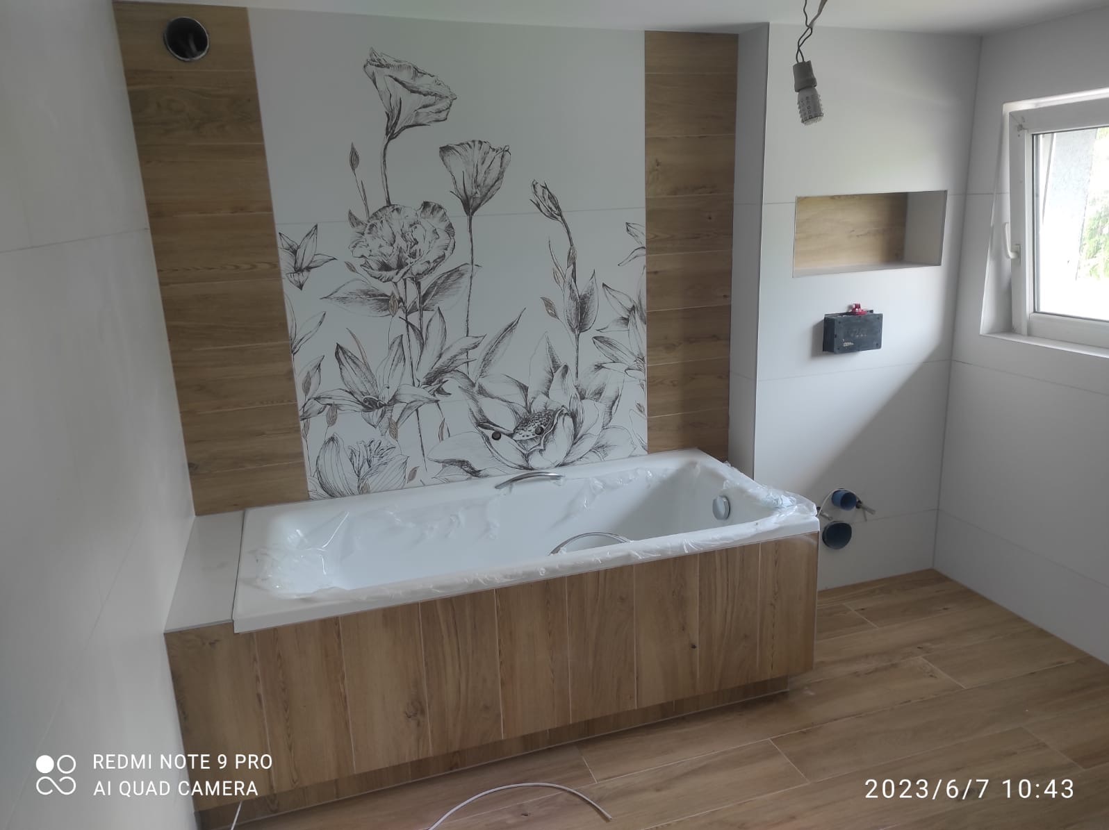 Wykończona łazienka z wanną obudowaną drewnopodobnymi panelami, płytki z motywem kwiatowym na ścianie, drewniana podłoga i wnęka w ścianie.