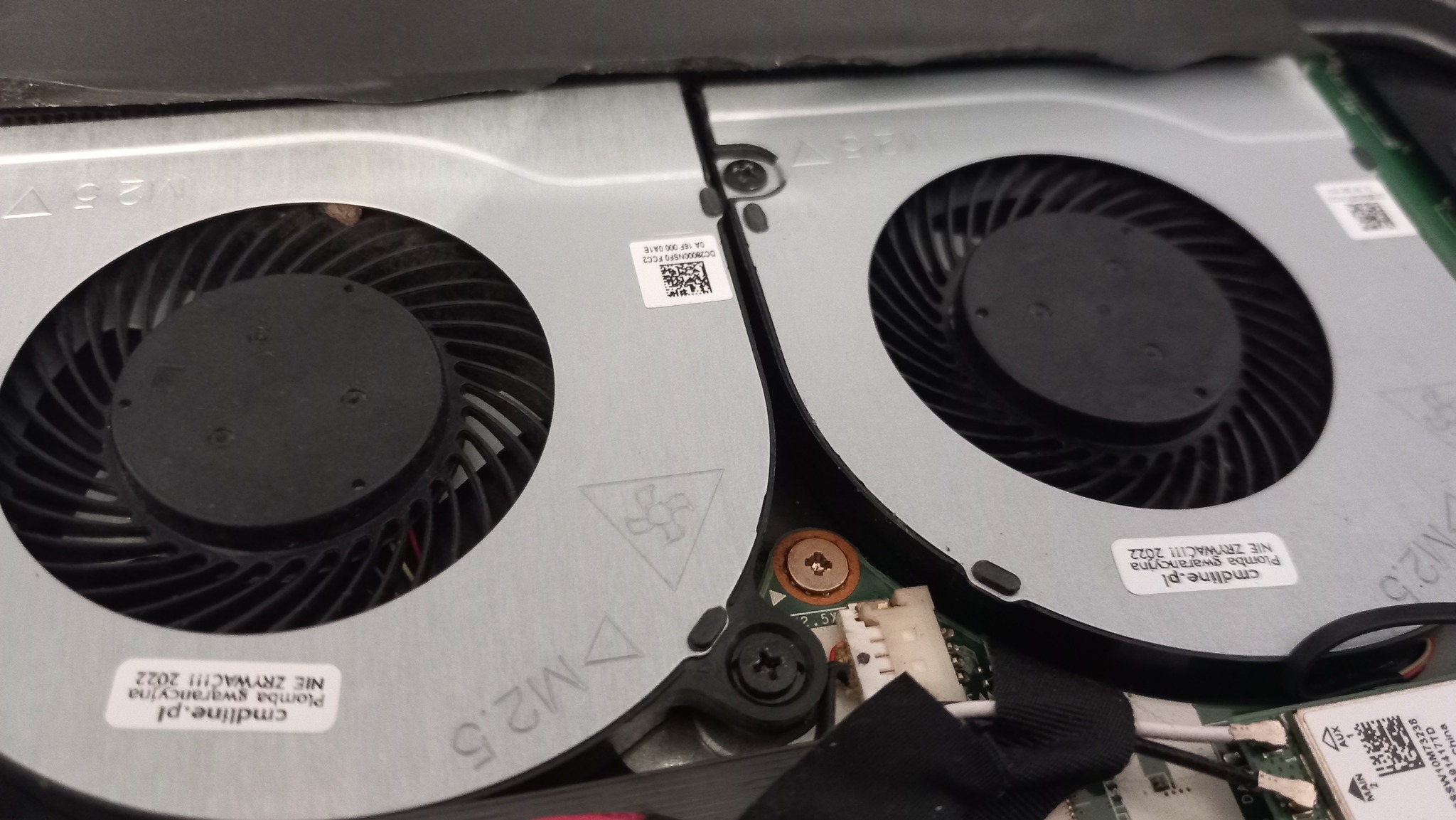 Wnętrze laptopa z widocznymi dwoma wentylatorami chłodzącymi, elementami płyty głównej i przewodami. Widoczne naklejki z informacjami o gwarancji i modelu.