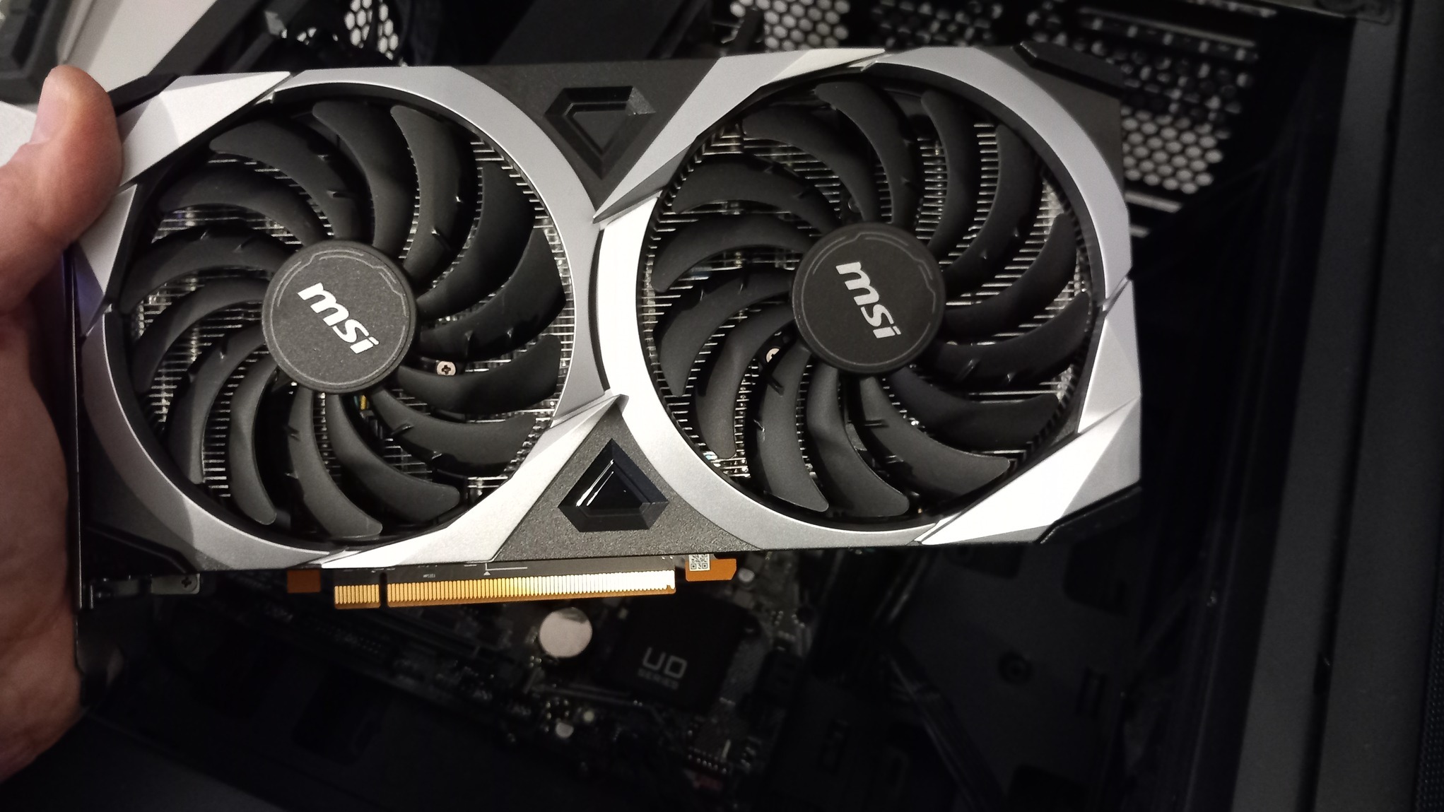 Karta graficzna MSI z dwoma wentylatorami, częściowo zamontowana w obudowie komputera. Widoczny fragment płyty głównej. Ujęcie z bliska, z ręką trzymającą kartę.