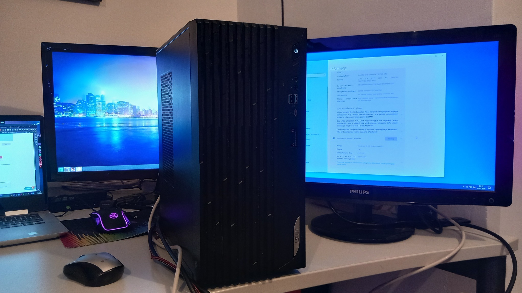 Czarna obudowa komputera stacjonarnego na biurku, widoczne dwa monitory, klawiatura i mysz. Na ekranie monitora otwarte okno z informacjami o systemie Windows.