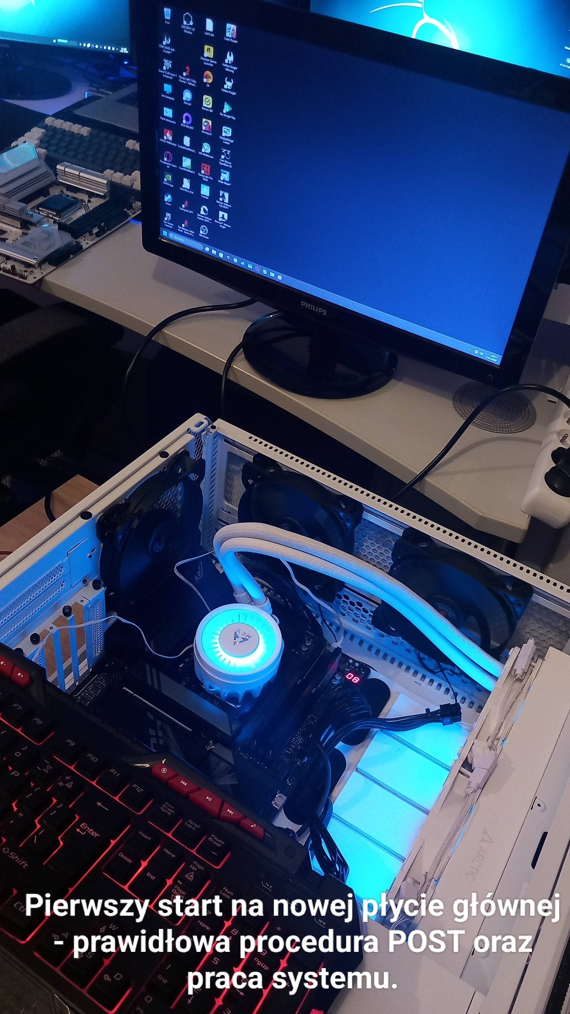 Komputer stacjonarny w trakcie montażu, widoczna płyta główna z chłodzeniem wodnym Arctic, monitor Philips i klawiatura. Test POST na nowej płycie głównej.