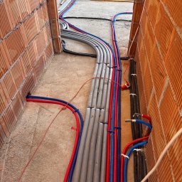 M&M instalacje - Instalacja rur (czerwone, niebieskie, szare) w bruzdach ceglanych ścian, widok z góry. Rury ułożone równolegle, zabezpieczone opaskami. W tle widoczne materiały budowlane.