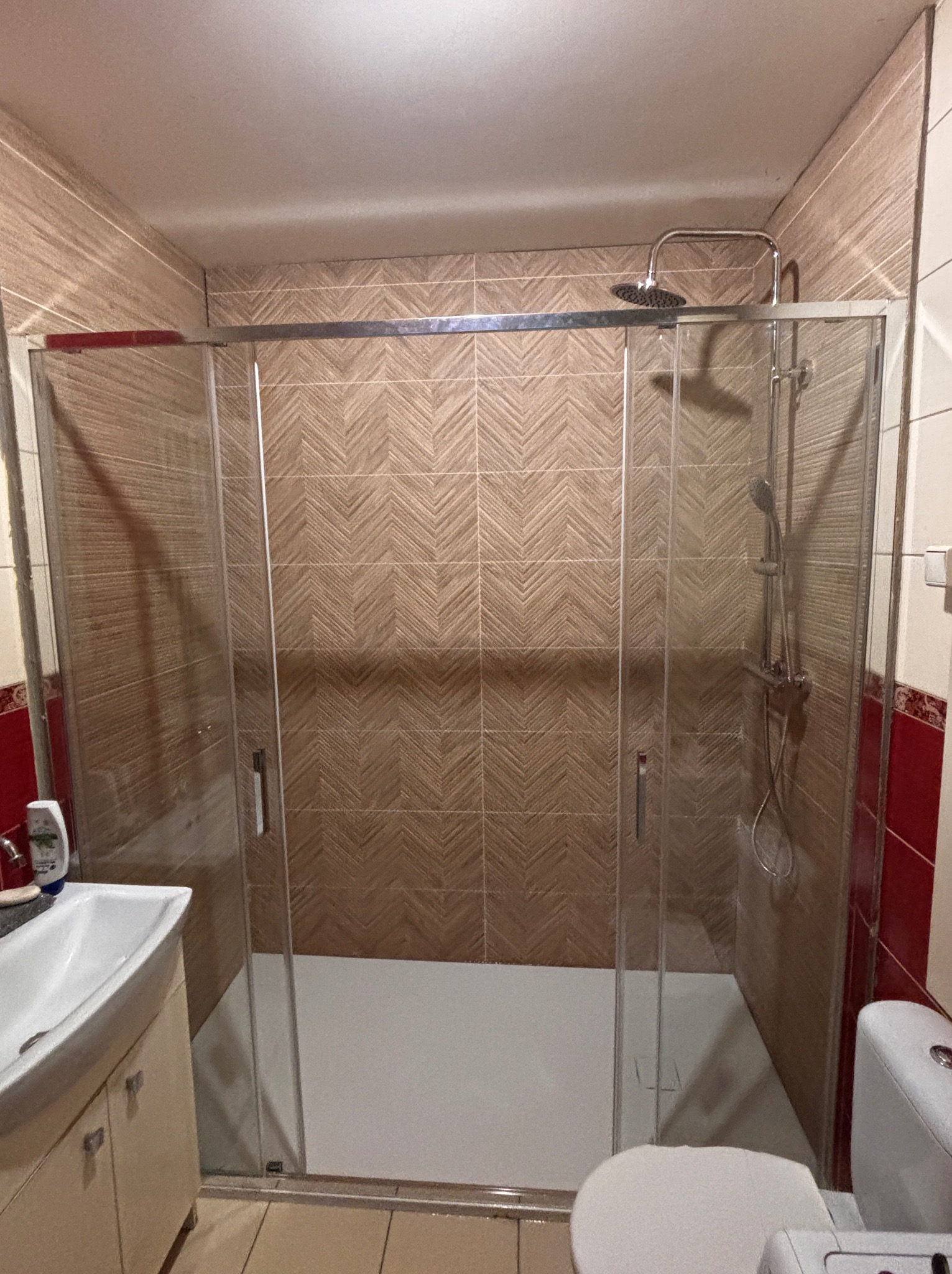 Nowoczesna kabina prysznicowa z deszczownicą, wykończona płytkami w odcieniach brązu i beżu. Widoczna umywalka z szafką i toaleta. Całość utrzymana w jasnej tonacji.