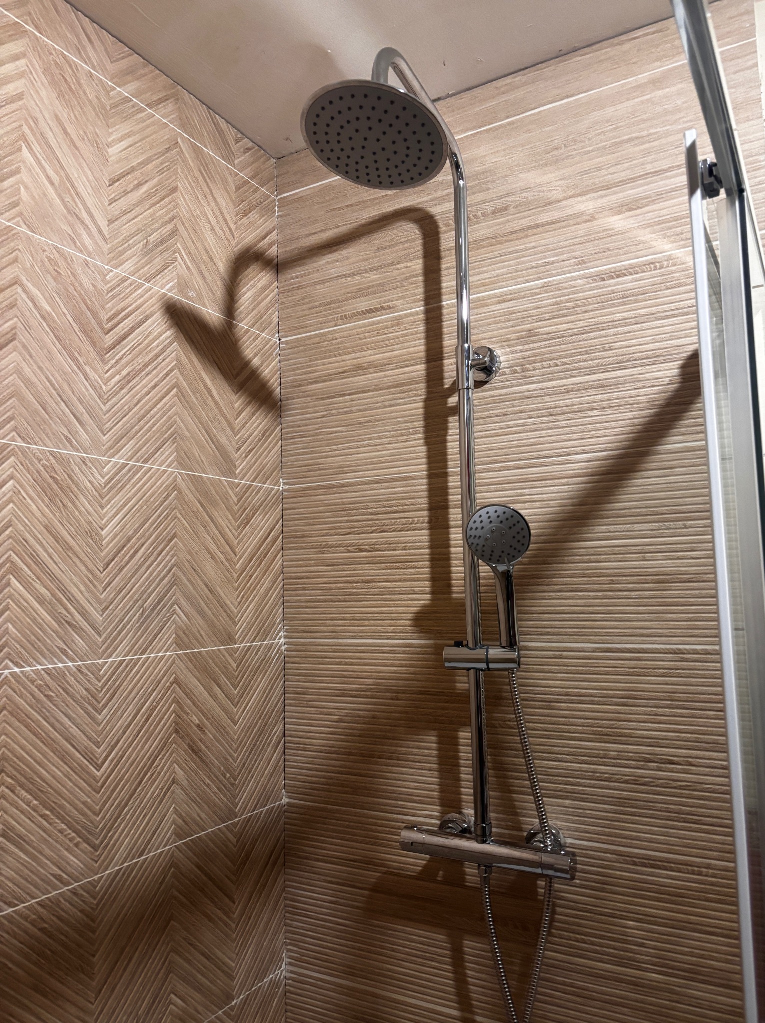 Nowoczesny zestaw prysznicowy z deszczownicą i słuchawką, zamontowany na ścianie z dekoracyjnymi płytkami w odcieniach brązu. Widoczna kabina prysznicowa.