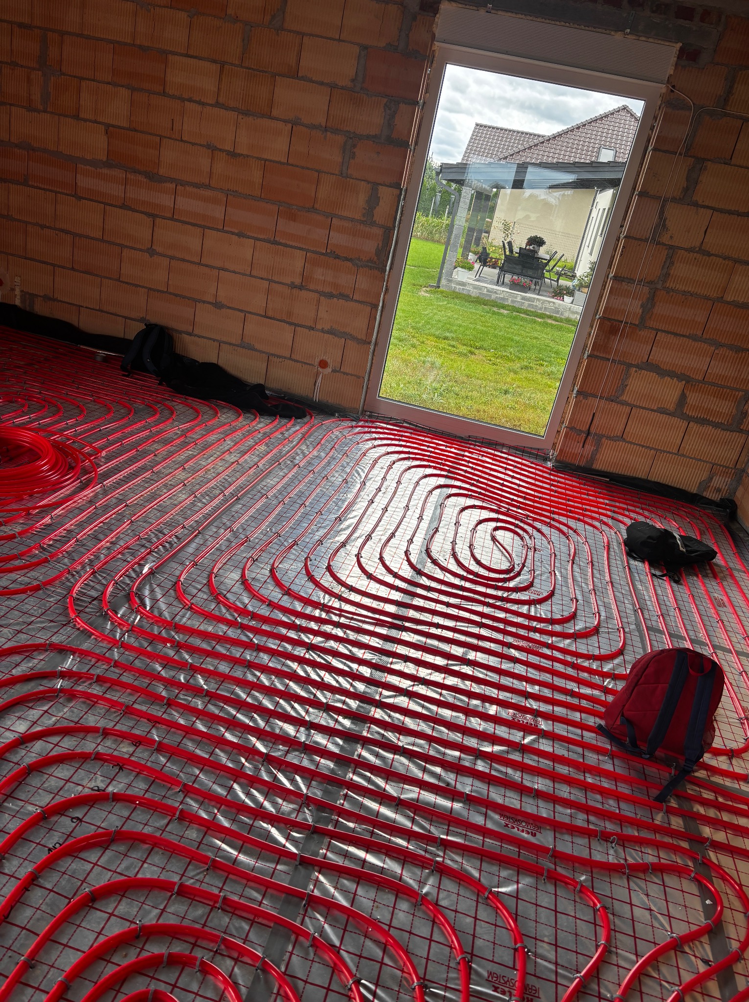 Czerwone rury ogrzewania podłogowego ułożone na folii w surowym wnętrzu z ceglanymi ścianami i widokiem na ogród przez okno. Torba robocza w rogu.