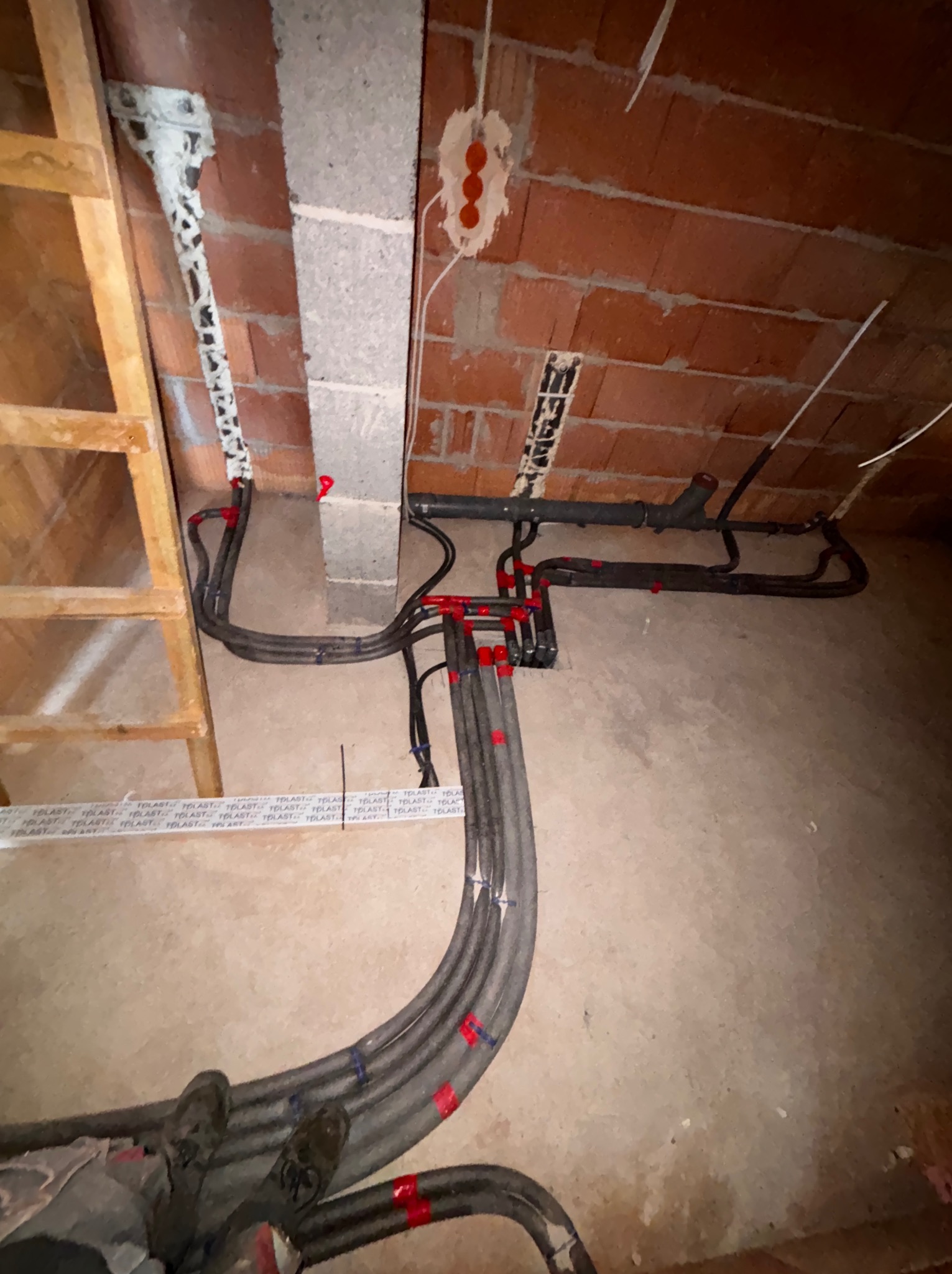 Szare rury instalacji grzewczej z czerwonymi oznaczeniami, ułożone na podłodze w surowym wnętrzu z ceglaną ścianą i widoczną instalacją elektryczną.