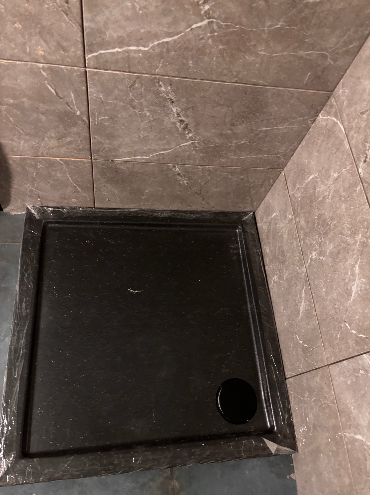 Czarny brodzik z odpływem, otoczony marmurowymi płytkami w odcieniach szarości. Widok z góry, ukazujący detale wykończenia i materiałów.