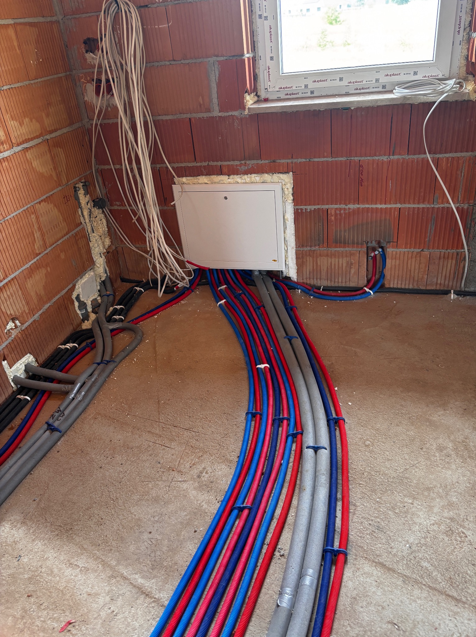 Instalacja wodna i elektryczna w surowym budynku: rury w kolorach czerwonym, niebieskim i szarym prowadzone do białej skrzynki rozdzielczej na ceglanej ścianie.