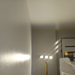 Michał - Świeżo pomalowana ściana z widocznymi smugami, oświetlona lampą budowlaną. Żółta taśma malarska zabezpiecza sufit. W tle drzwi i fragment lampy.