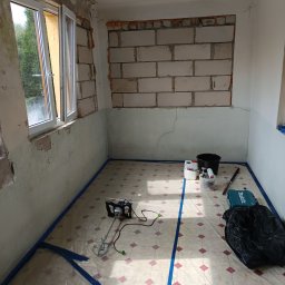 Michał - Pokój w trakcie remontu: okno, ściany z ubytkami, zabezpieczona podłoga folią i taśmą. Widoczne narzędzia i materiały przygotowane do szpachlowania.