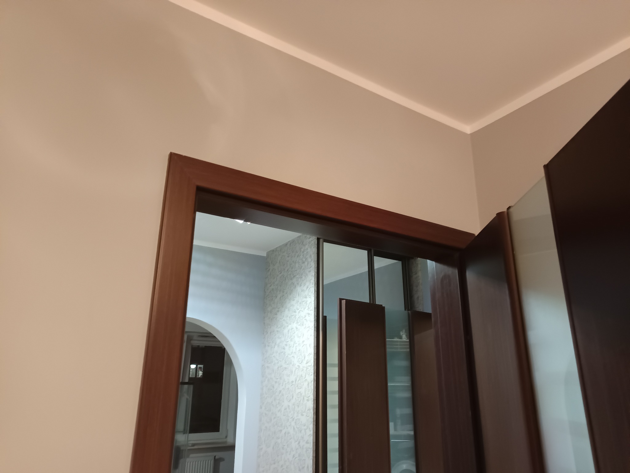 Fragment wykończonego wnętrza: brązowa rama drzwiowa, pomalowana ściana w odcieniach beżu i widok na pokój z tapetą w delikatny wzór. Widoczny fragment sufitu z listwą.