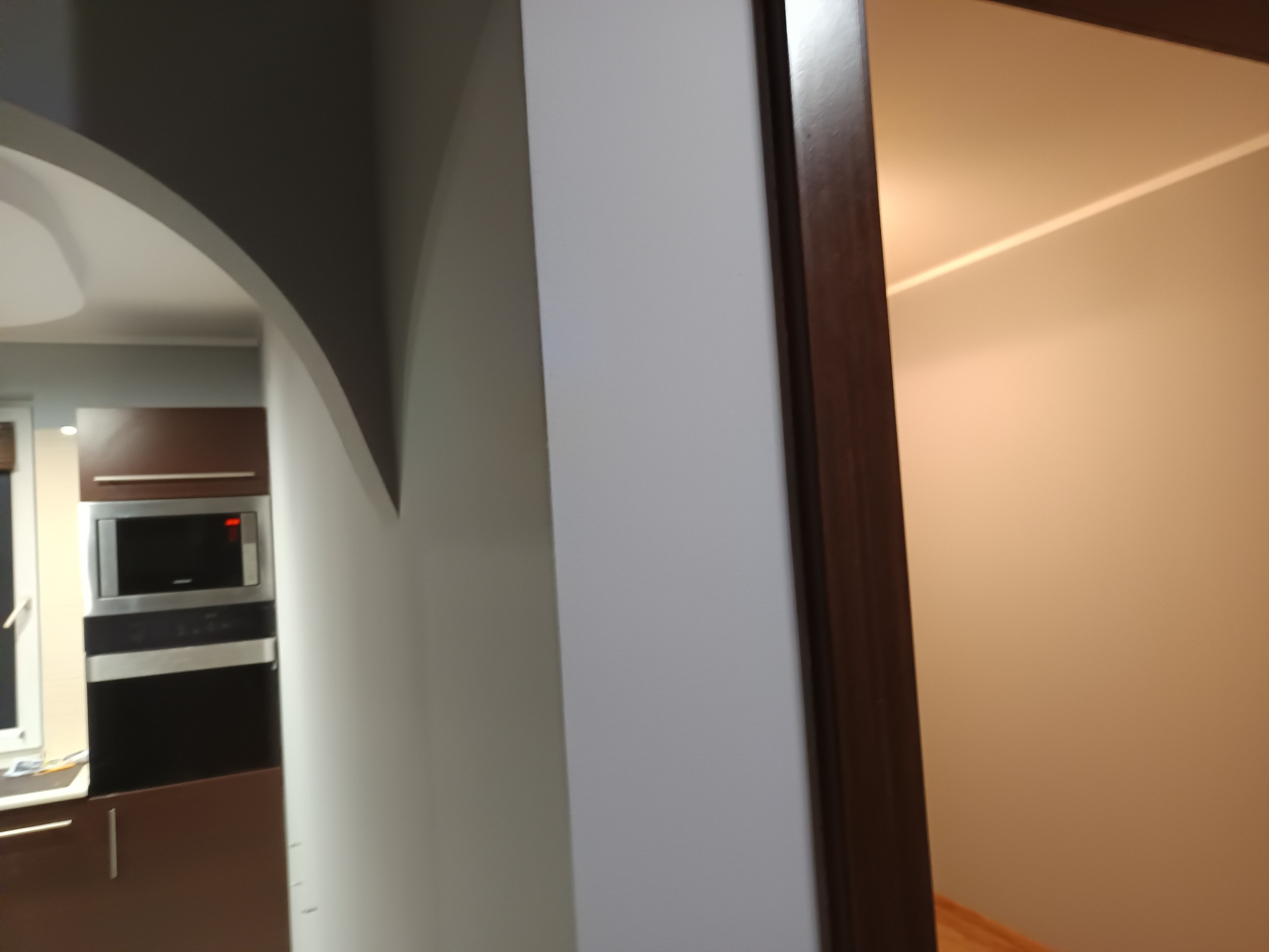 Nowoczesna kuchnia z łukowym przejściem do salonu, widok z perspektywy korytarza. Minimalistyczny design, brązowe szafki i białe ściany.