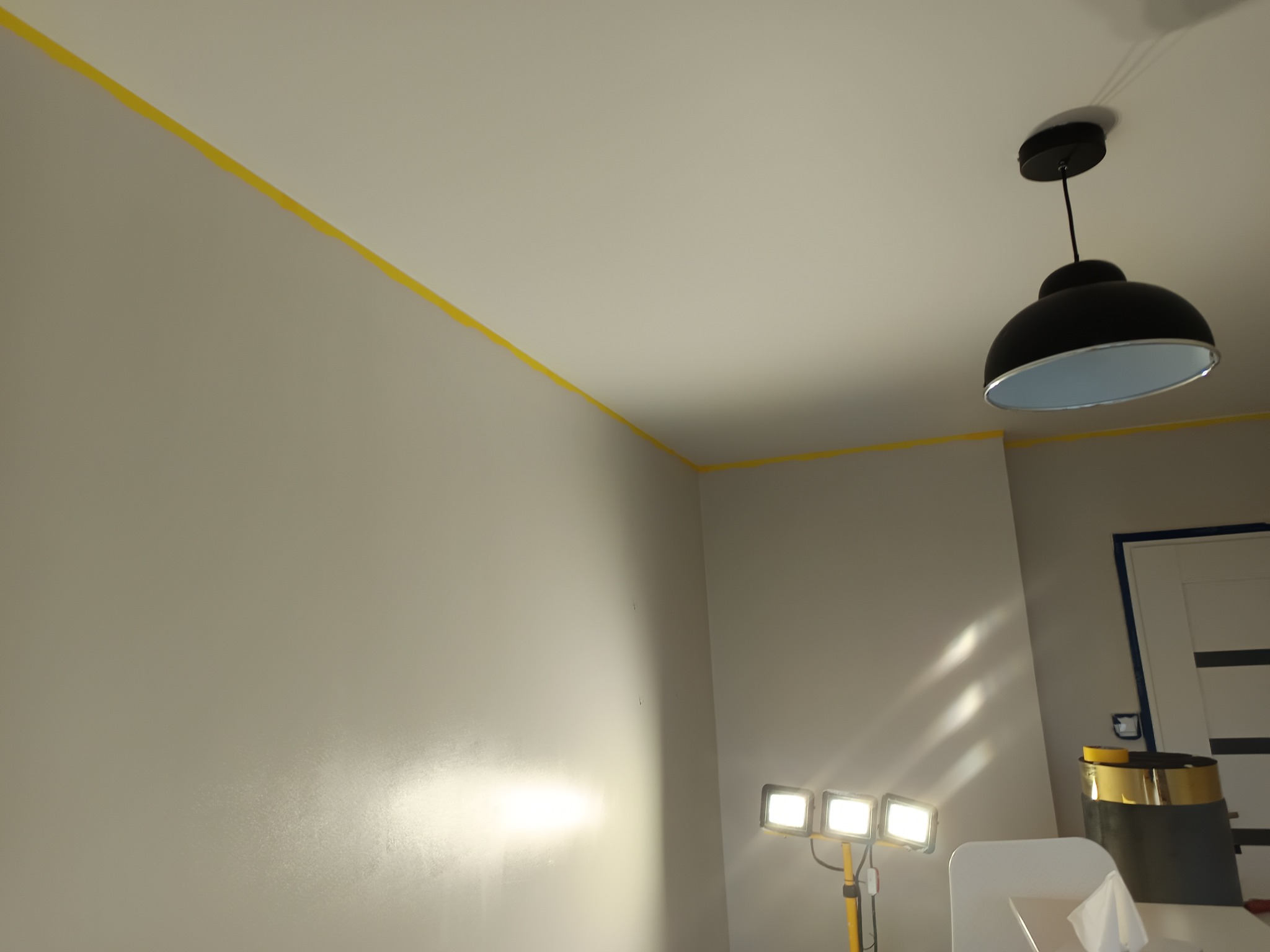 Ściana w trakcie malowania, zabezpieczona żółtą taśmą malarską przy suficie. Widoczne oświetlenie robocze i lampa sufitowa. Przygotowanie do malowania.