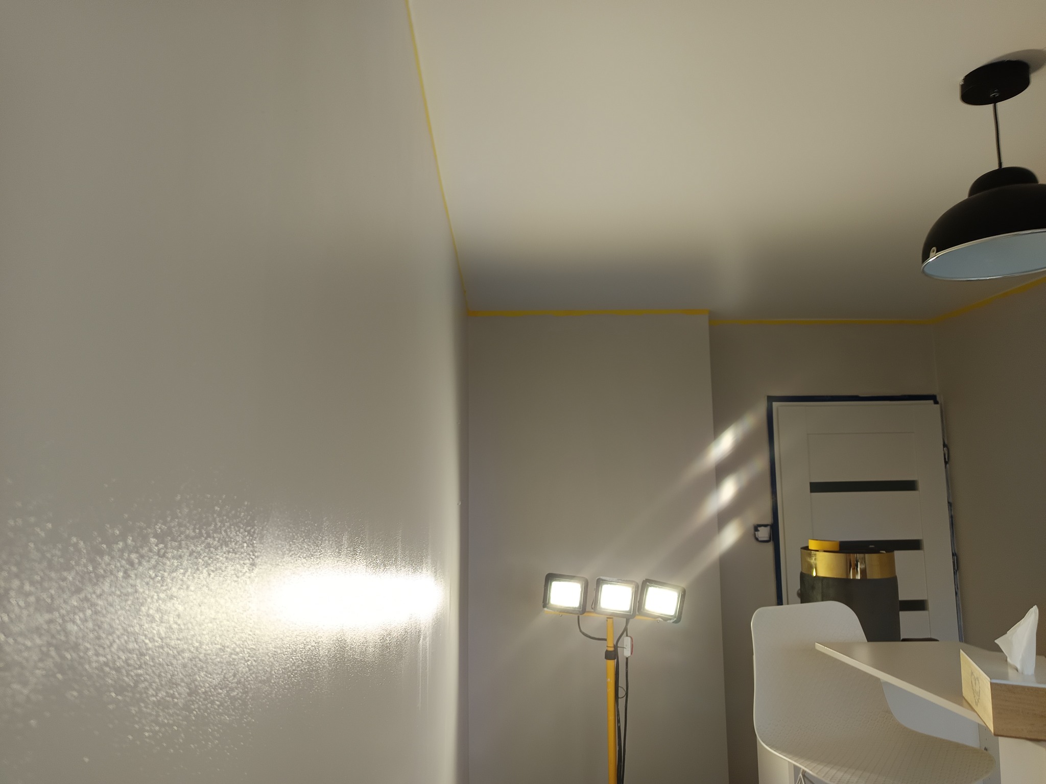 Świeżo pomalowana ściana z widocznymi smugami, oświetlona lampą budowlaną. Żółta taśma malarska zabezpiecza sufit. W tle drzwi i fragment lampy.