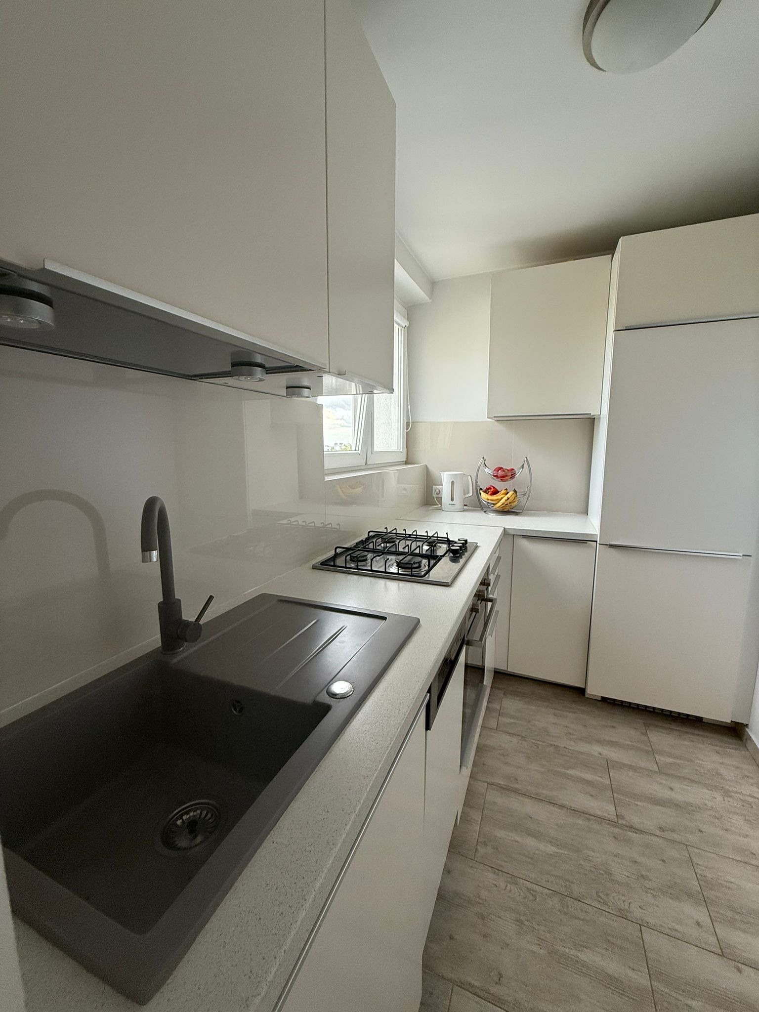 Jasna kuchnia po remoncie: minimalistyczne białe szafki, szary zlew z baterią, płyta gazowa i drewnopodobne płytki podłogowe. Nowoczesne wykończenie wnętrza.