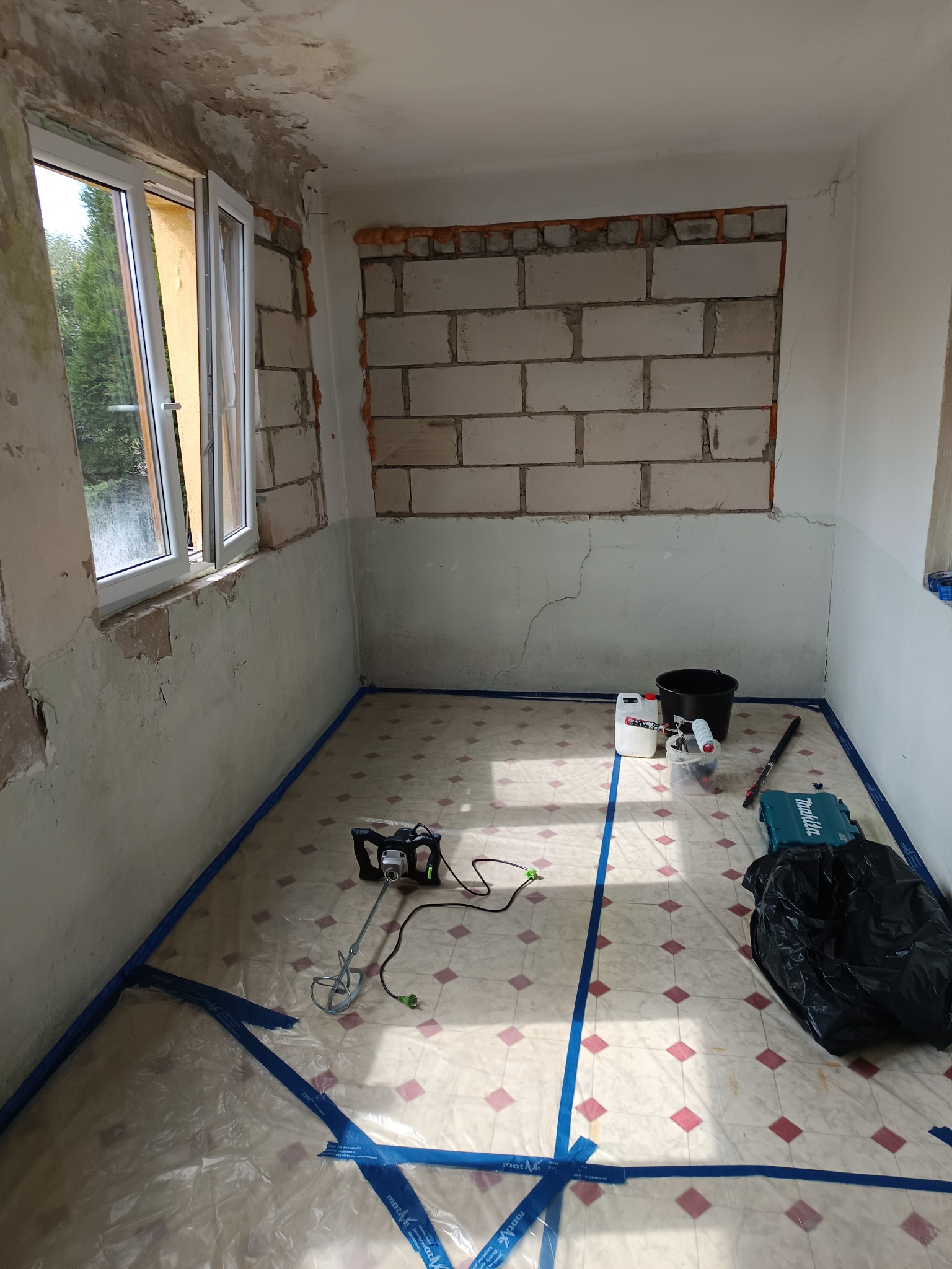 Pokój w trakcie remontu: okno, ściany z ubytkami, zabezpieczona podłoga folią i taśmą. Widoczne narzędzia i materiały przygotowane do szpachlowania.