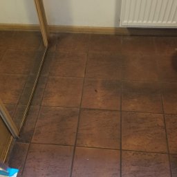 Royal Clean Studio Żaneta Spała - Brudna, brązowa podłoga z płytek ceramicznych w pomieszczeniu, widoczny fragment drabiny i grzejnika. Wymaga sprzątania.