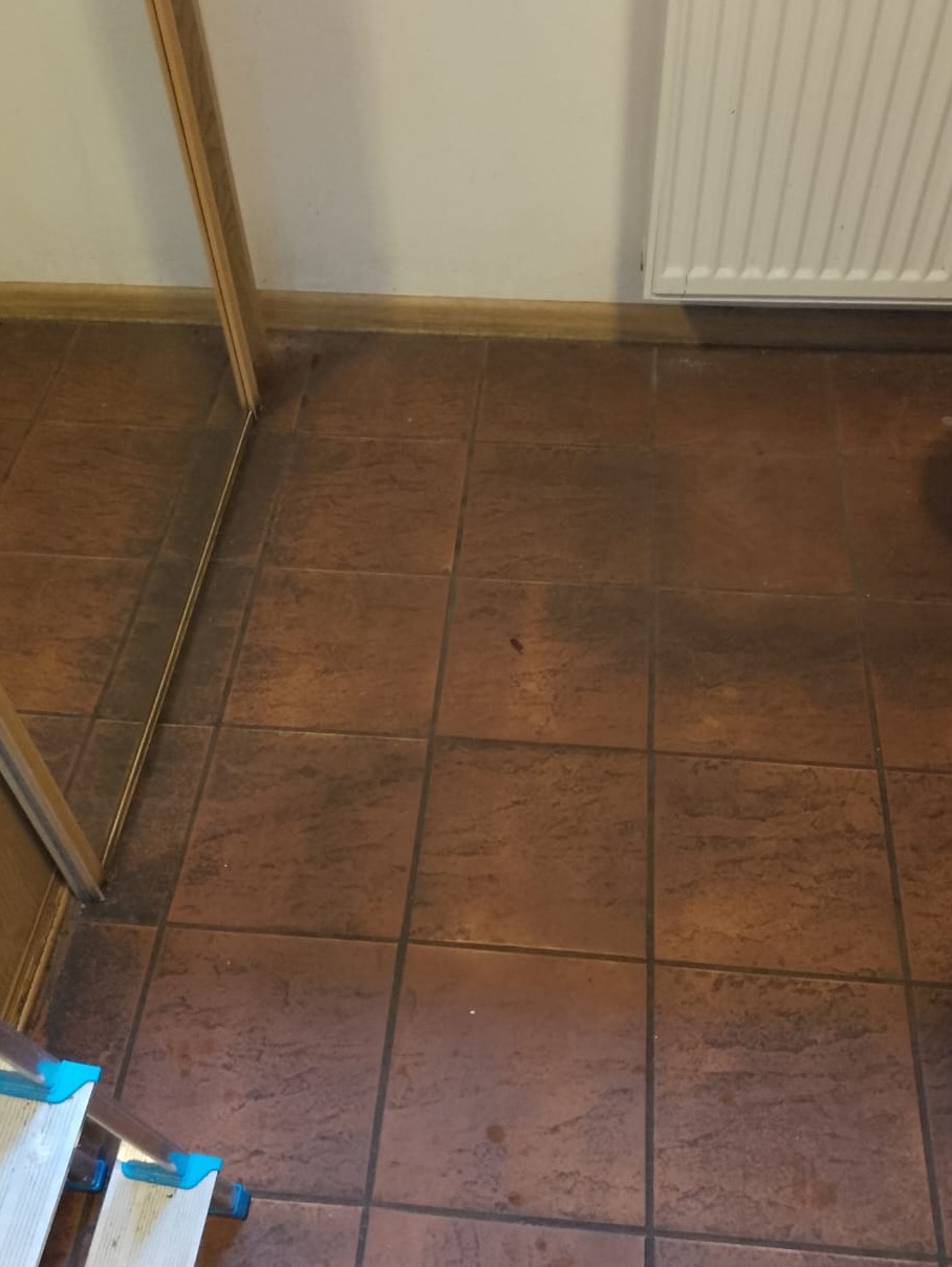 Brudna, brązowa podłoga z płytek ceramicznych w pomieszczeniu, widoczny fragment drabiny i grzejnika. Wymaga sprzątania.