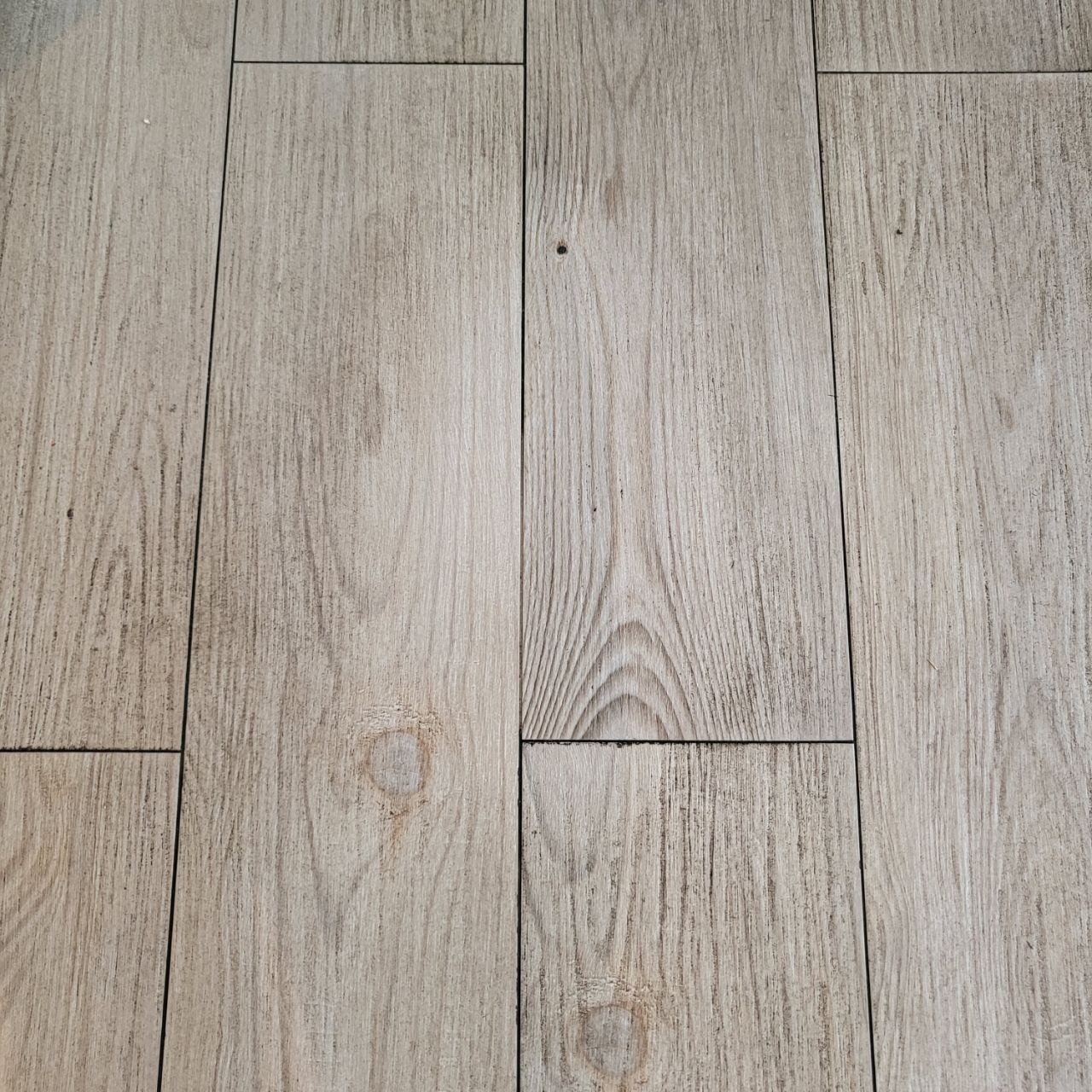 Jasne, drewnopodobne panele podłogowe ułożone w regularny wzór, z widocznymi słojami i delikatnymi zabrudzeniami w fugach. Widok z góry.