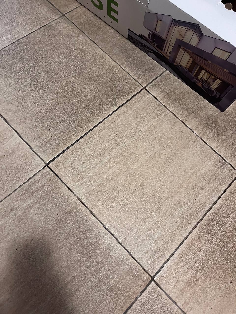 Beżowe płytki podłogowe z ciemnymi fugami, fragment opakowania z wizualizacją nowoczesnego domu w prawym górnym rogu. Ujęcie z góry, lekki kąt.