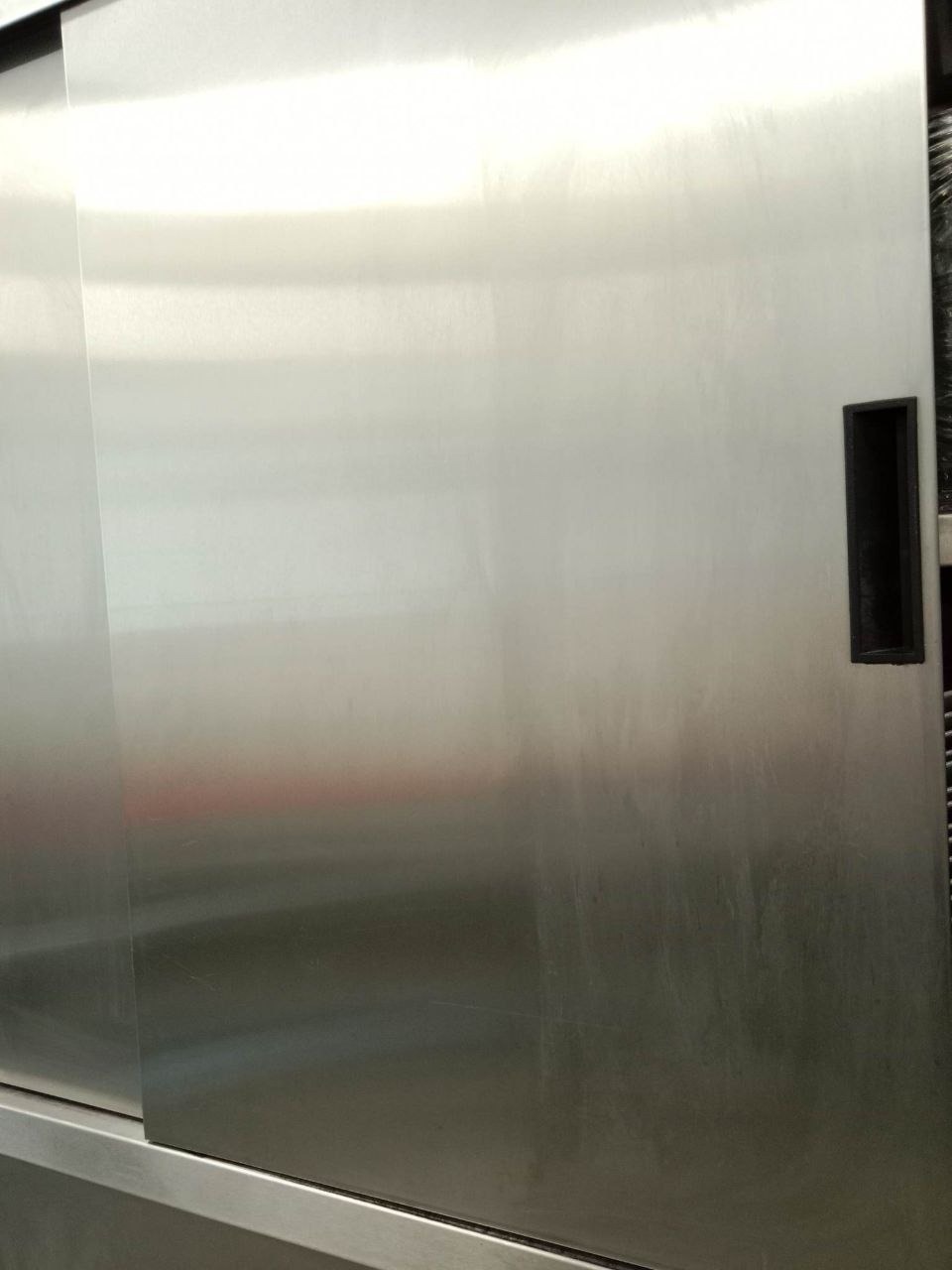 Błyszcząca, metalowa powierzchnia przesuwnych drzwi z czarnym uchwytem, widoczne smugi i niedoskonałości po czyszczeniu, bliski kadr.