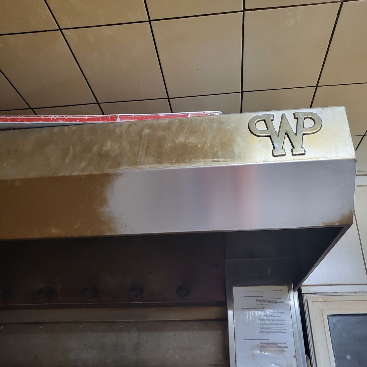 Brudny okap kuchenny ze stali nierdzewnej z logo 'WP' na tle beżowych płytek. Widoczne ślady tłuszczu i niedoczyszczone powierzchnie.