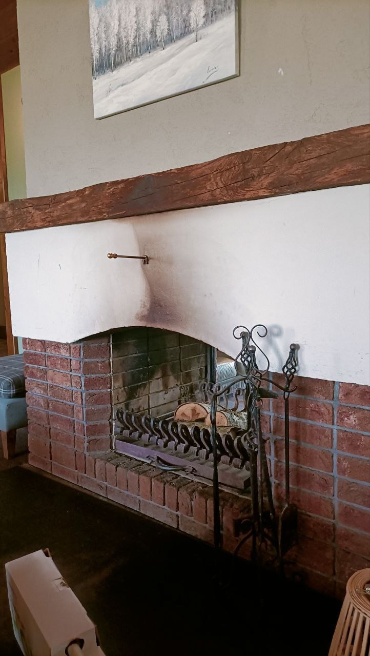 Brudny kominek z cegły i drewna z śladami sadzy na ścianie, przed czyszczeniem. Nad kominkiem obraz z zimowym krajobrazem.